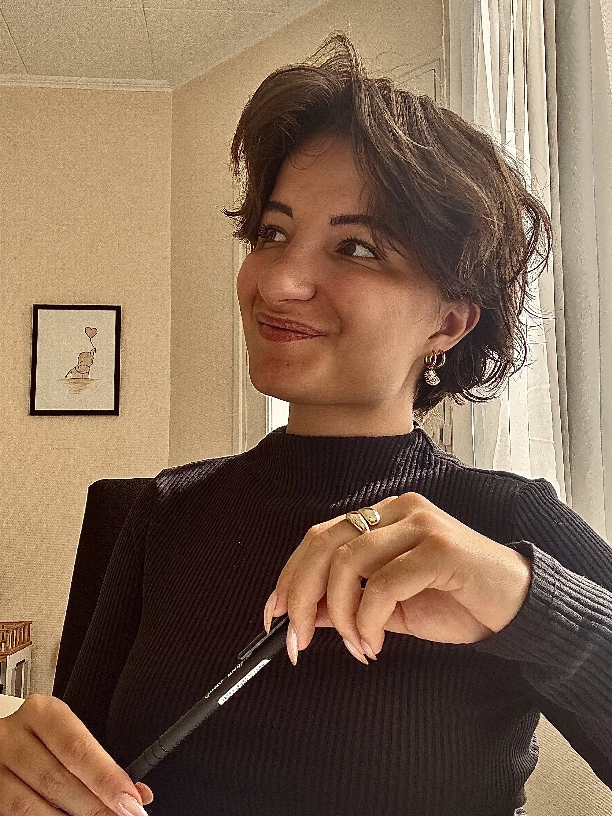 Une femme souriante aux cheveux courts, portant des boucles d'oreilles et un pull noir, assise à un bureau à côté d'une fenêtre avec des rideaux blancs, tenant un stylo dans la main.