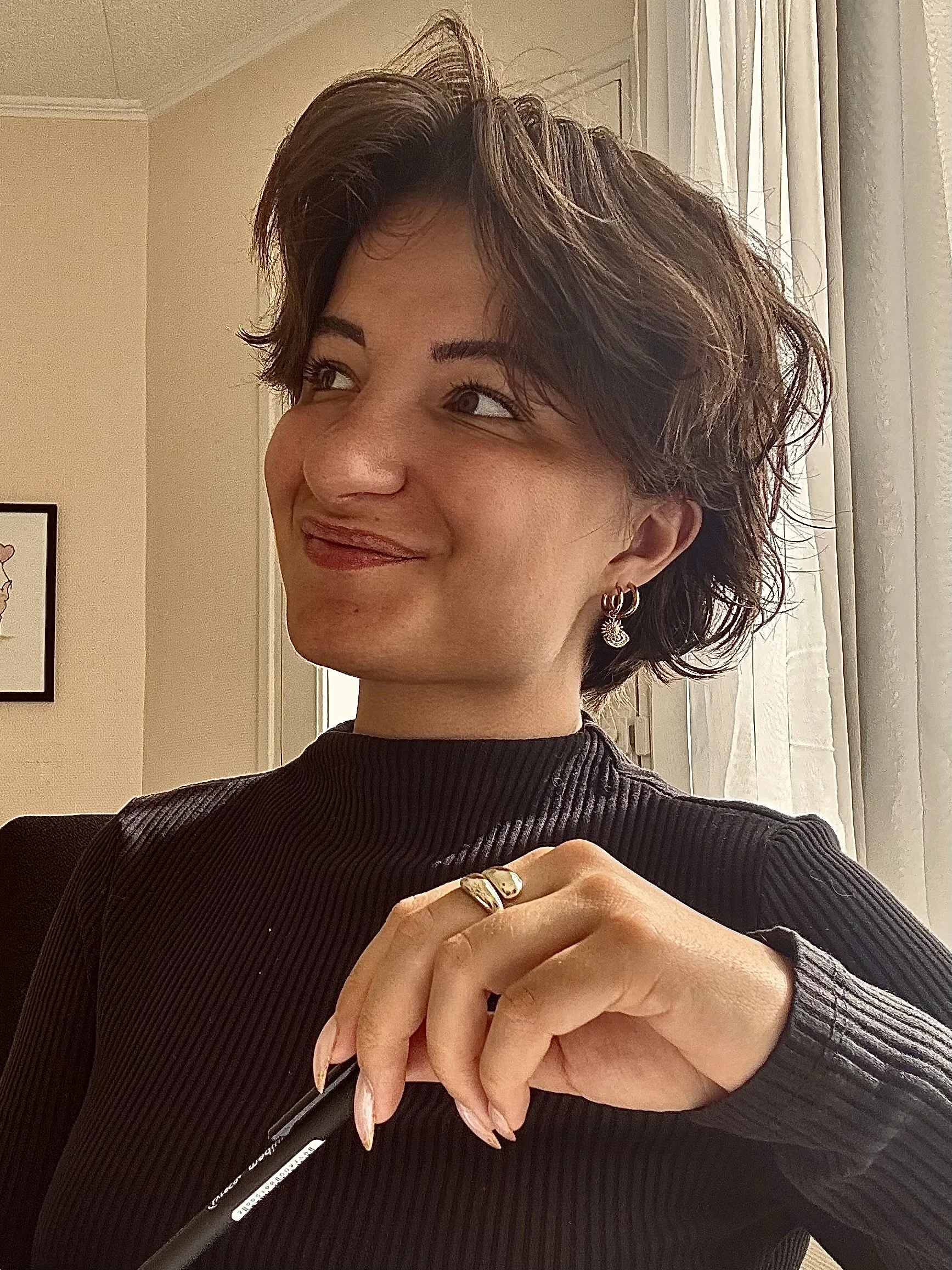 Une femme souriante avec des cheveux courts, portant un pull noir à col roumain et des boucles d'oreilles, tenant un stylo dans sa main. Elle est assise près d'une fenêtre avec des rideaux blancs, dans une pièce lumineuse.