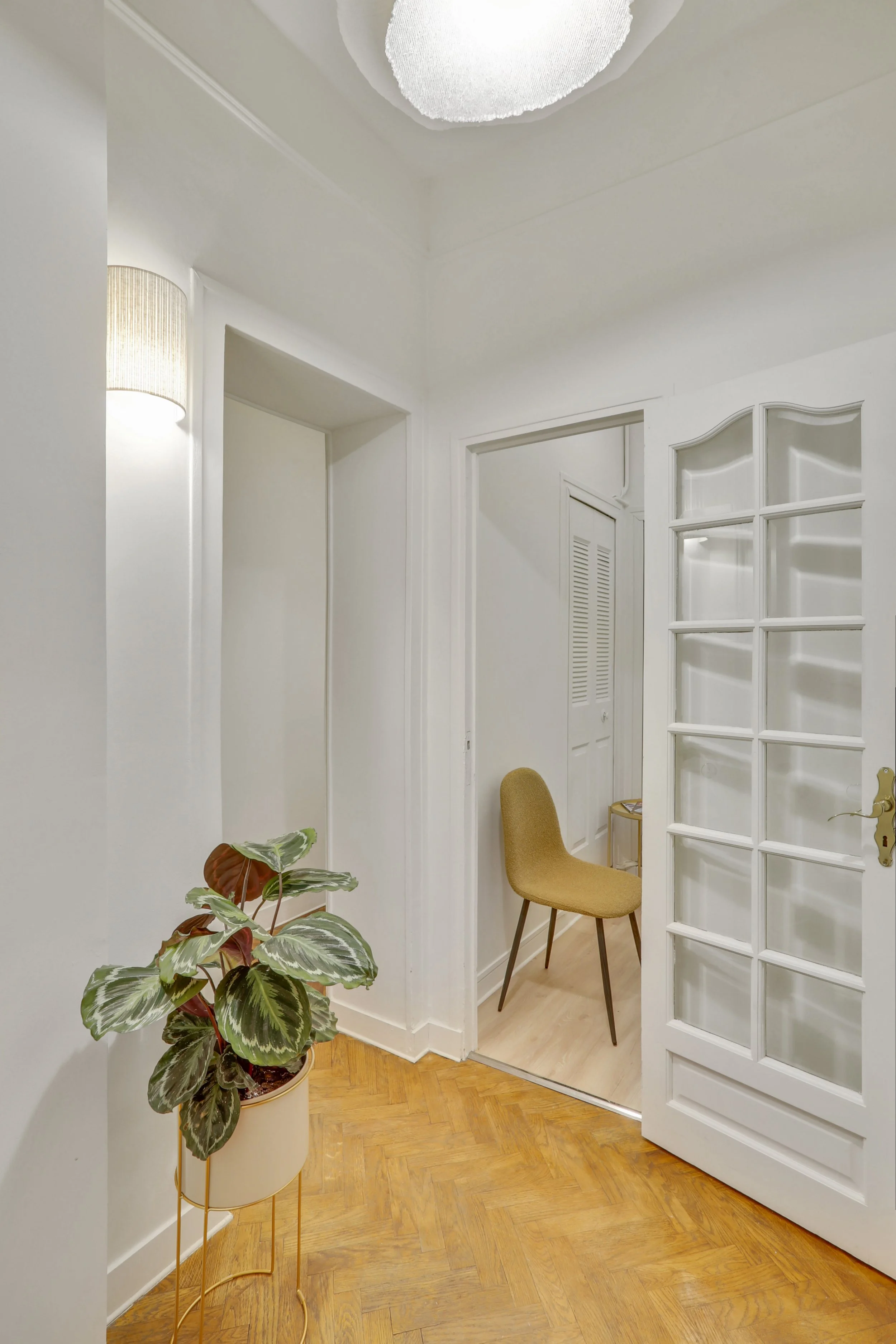 Intérieur d'une pièce avec un mur blanc, une porte en verre, un fauteuil beige, une plante verte en pot, un sol en parquet