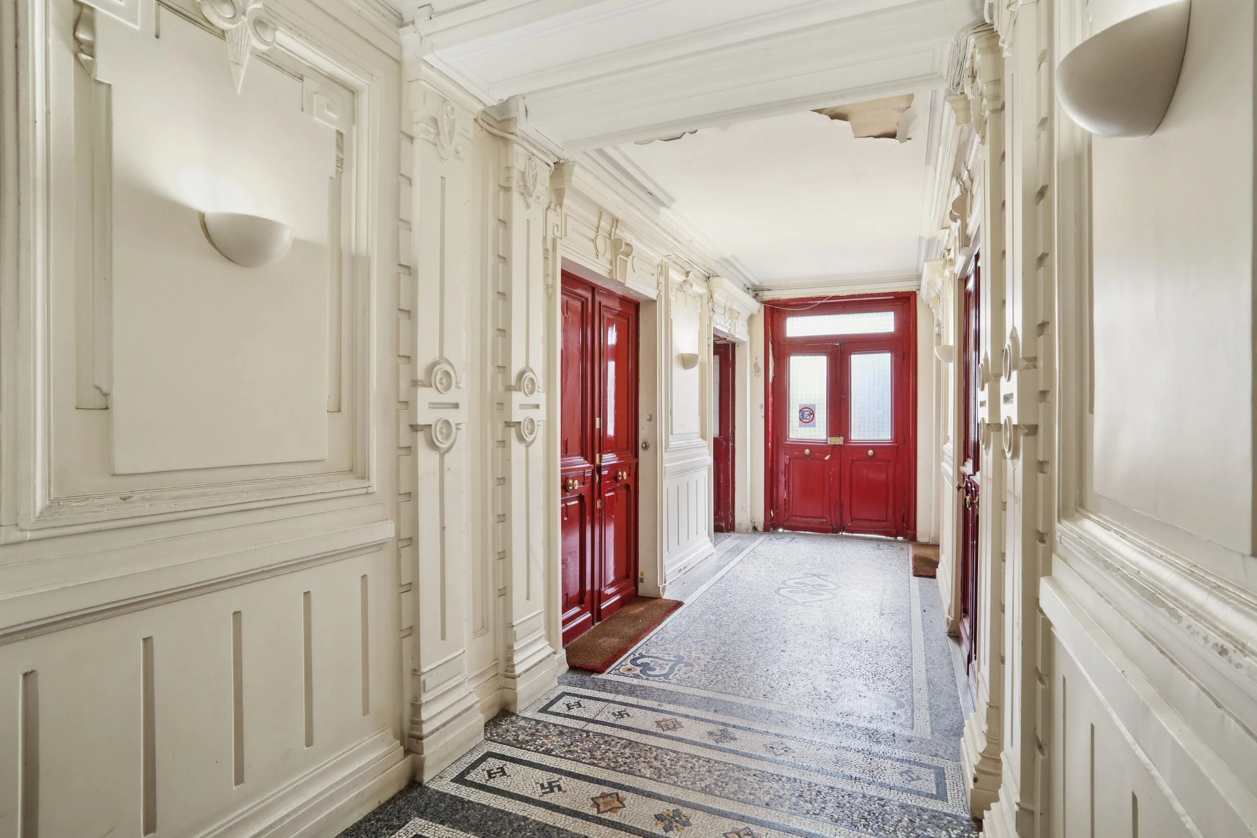 Entrée d'un immeuble ancien avec portes en bois rouge et sol en mosaïque, murs ornés de moulures blanches.