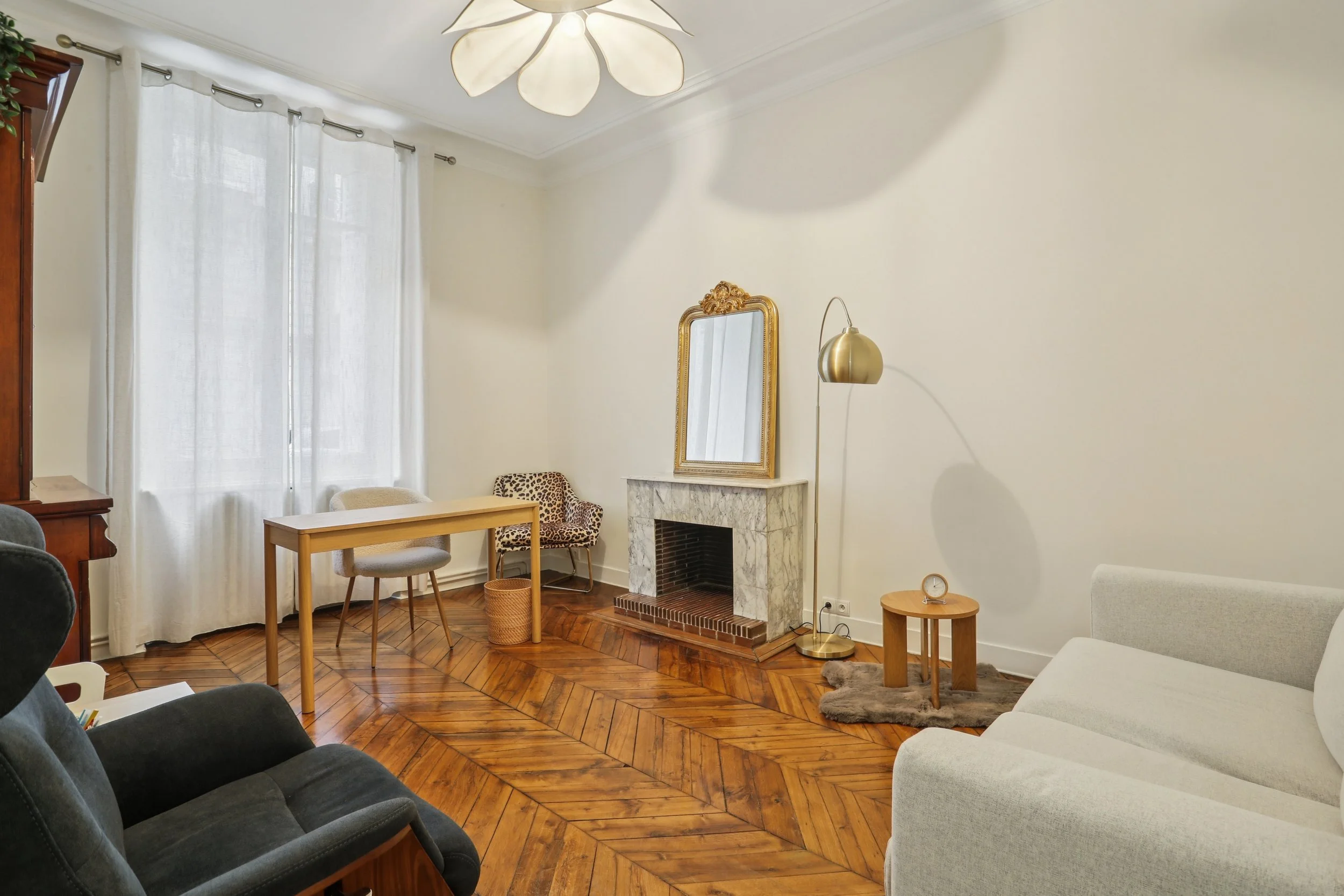 Salon avec cheminée en marbre, miroir doré au-dessus, canapé blanc, fauteuils variés, grande fenêtre avec rideaux blancs, parquet en bois, lampe de lecture dorée, tapis en fourrure