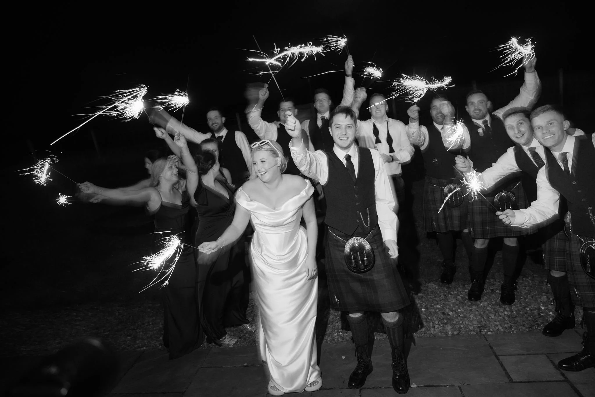 People celebrating at a night event, holding sparklers, with a woman in a white gown and a man in a kilt in the foreground.