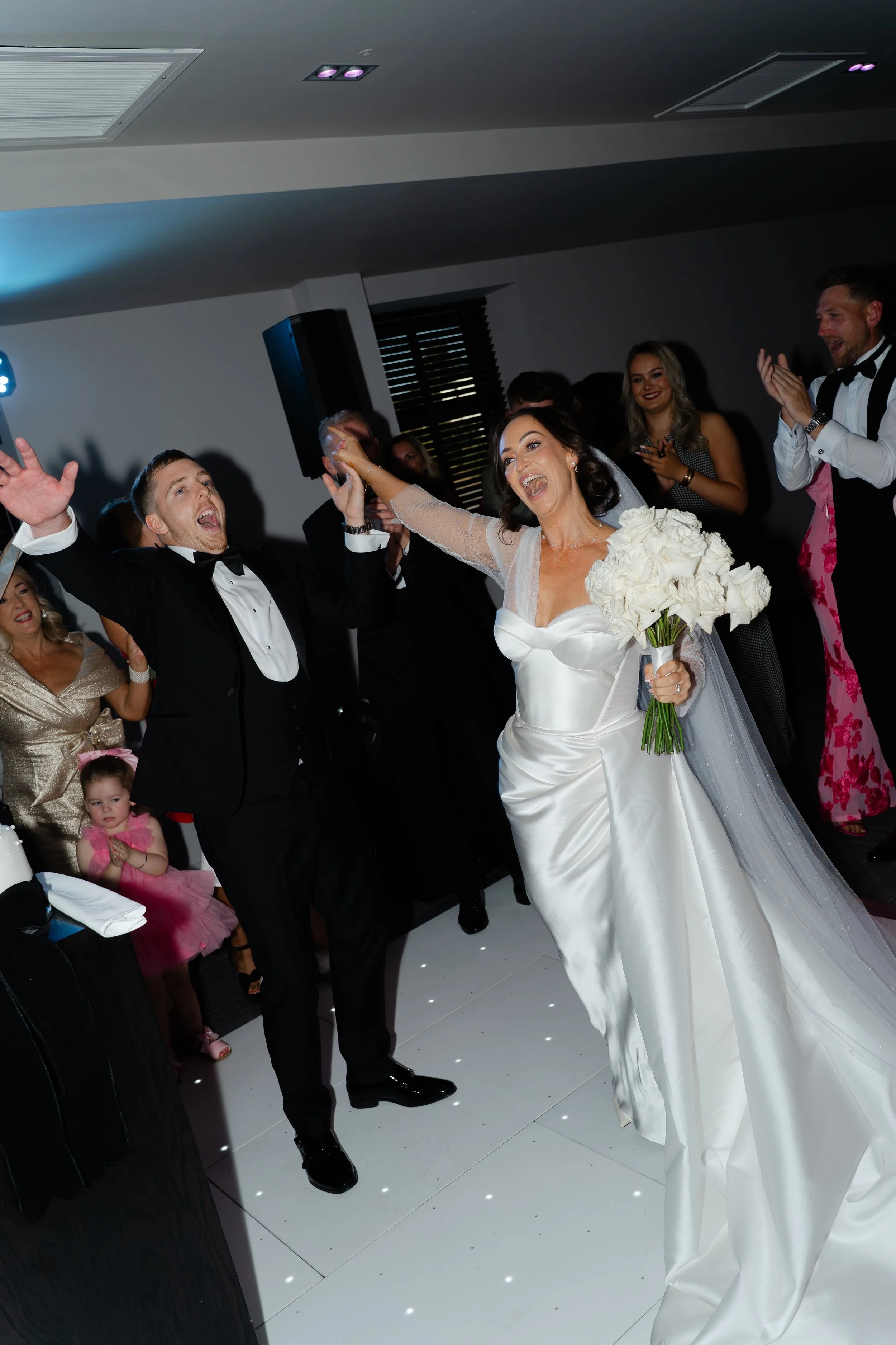 Bride and groom celebrating on the dance floor at their wedding reception, surrounded by guests clapping and cheering.