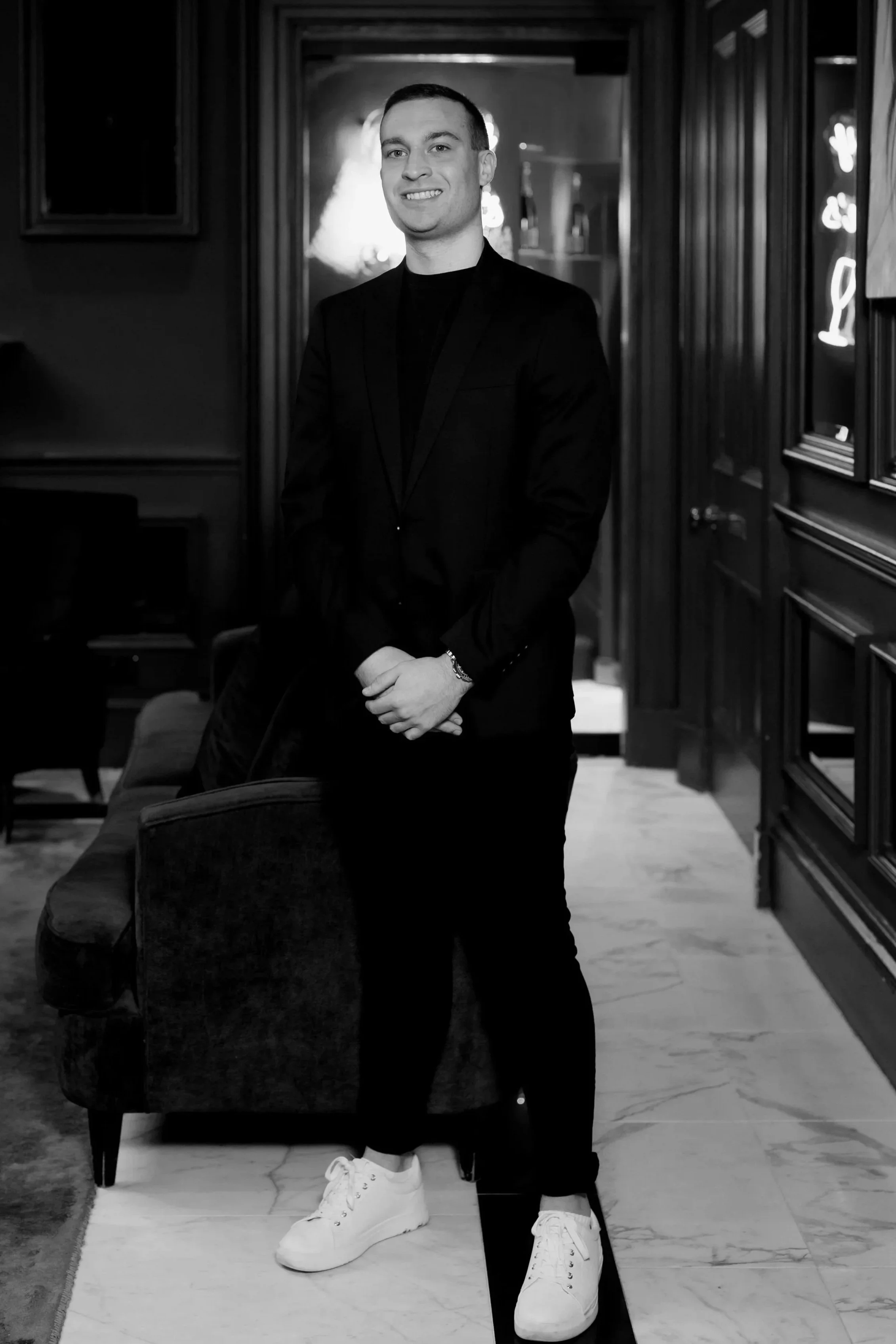 A young man in a black suit and white sneakers, smiling, standing in a dimly lit, elegant interior with dark wood paneling and framed art, with his hands clasped in front of him.