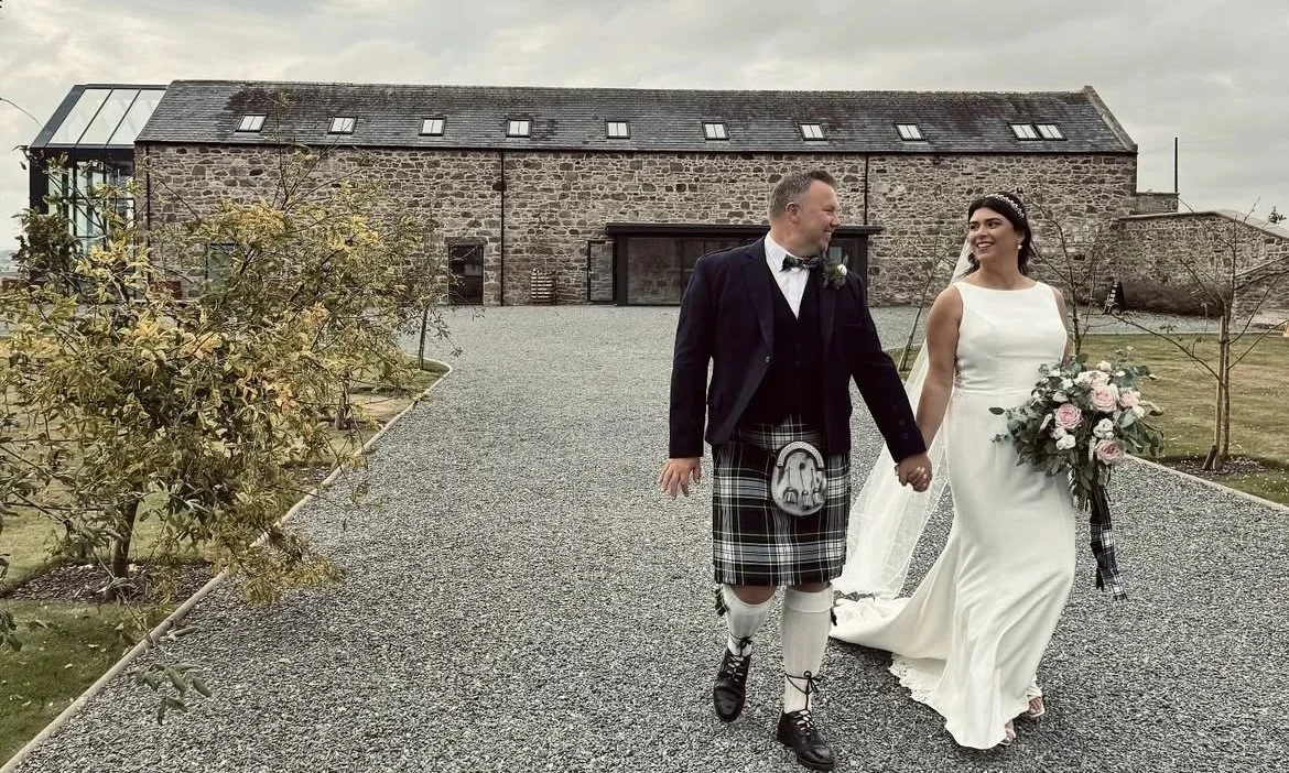 A bride and groom walking outdoors on a gravel path, holding hands, with a historic stone building in the background. The groom is wearing traditional Scottish attire, including a kilt, and the bride is in a white wedding dress holding a bouquet of flowers.
