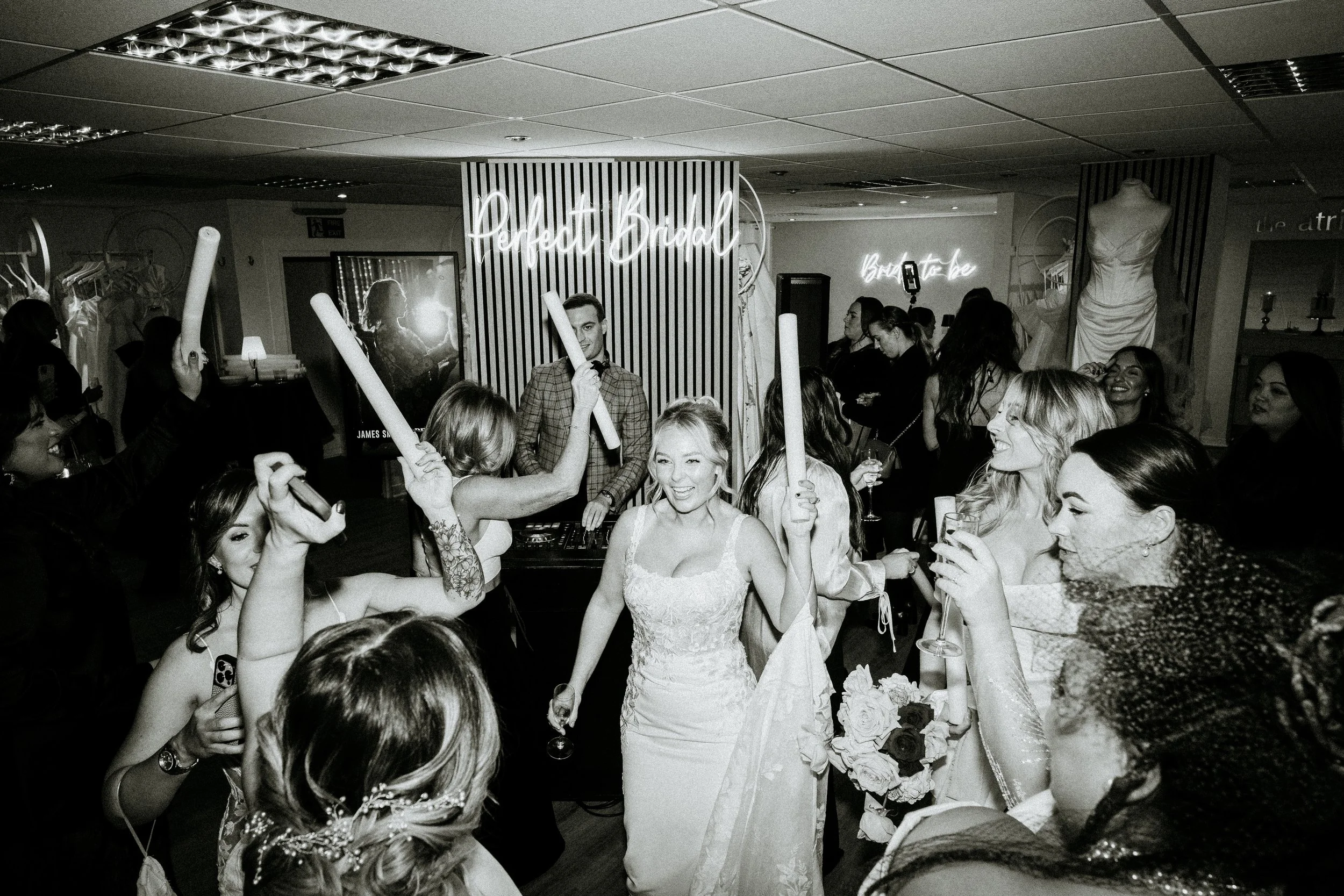 A bride in a wedding dress holding a bouquet smiles as she dances with guests at her wedding celebration. Guests are holding rolled napkins and drinking glasses, and others take photos. There is a neon sign that says 'Perfect Bridal' behind them, and a DJ in the background.