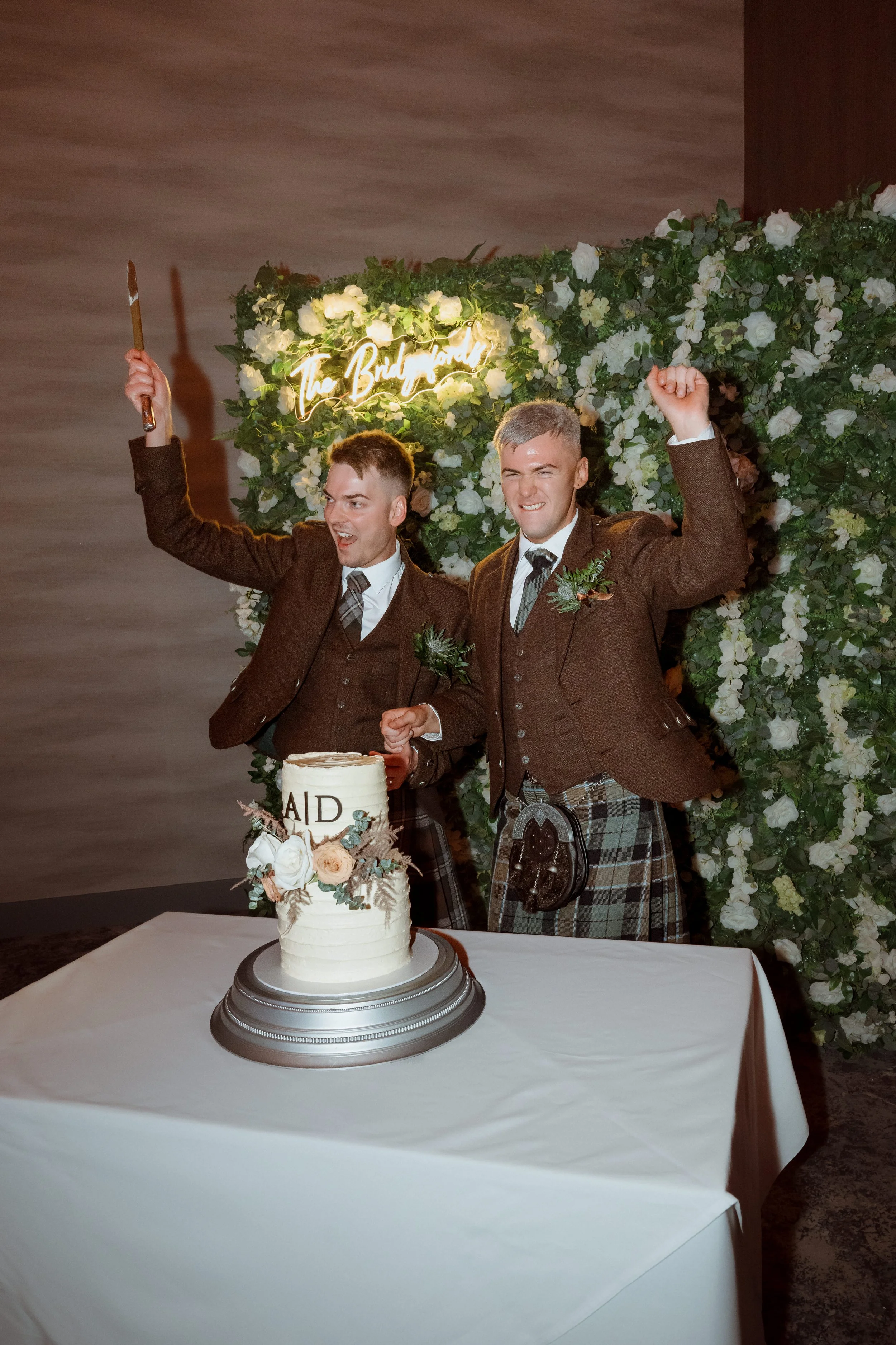 Two men dressed in brown suits and kilts celebrating behind a cake at a wedding or special event, with a floral backdrop and neon sign reading 'The Bridal Lounge'.