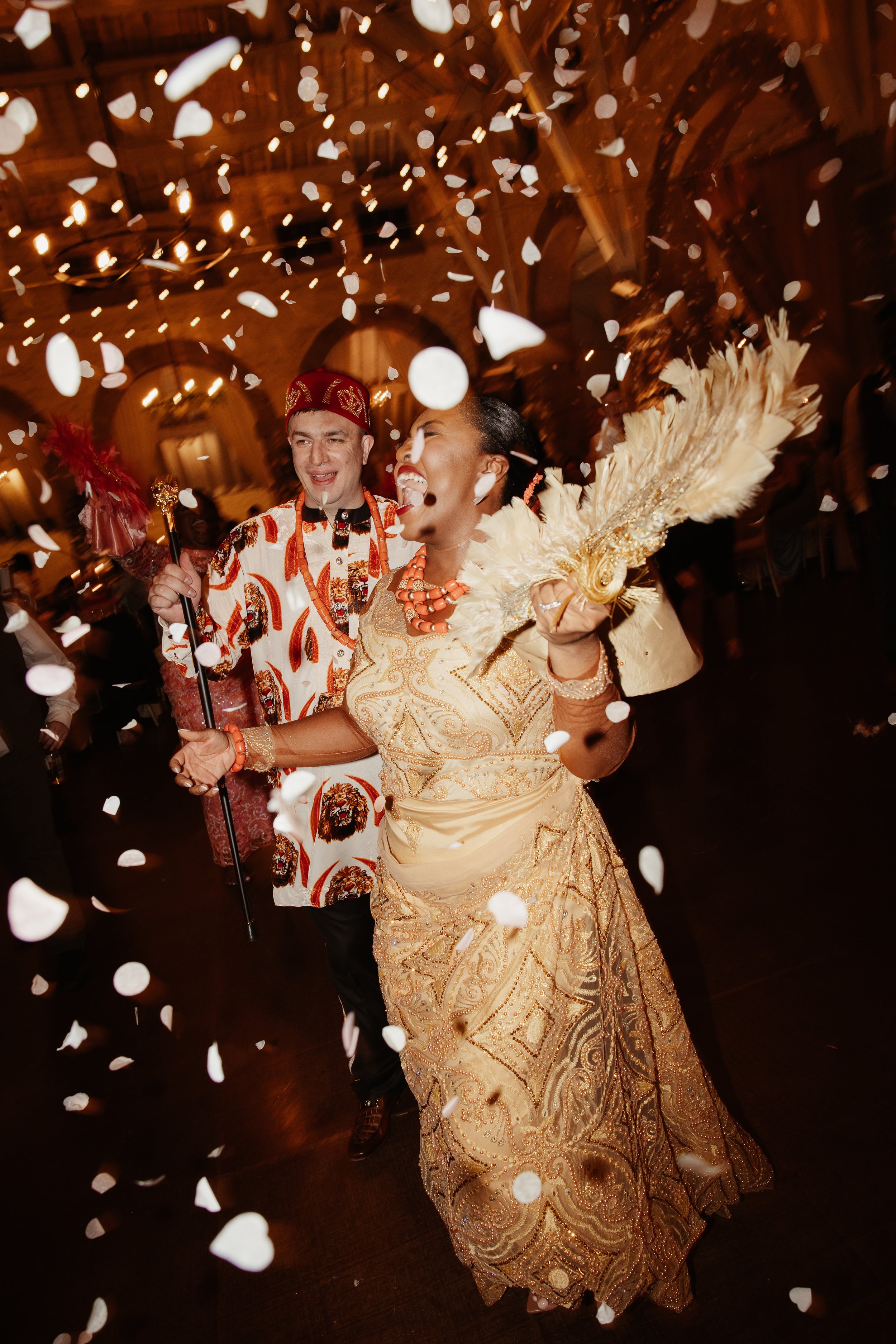 Couple celebrating at a wedding with confetti, the woman wearing traditional Indonesian attire with a fan, and the man in a patterned shirt and hat, dancing in a decorated indoor venue.