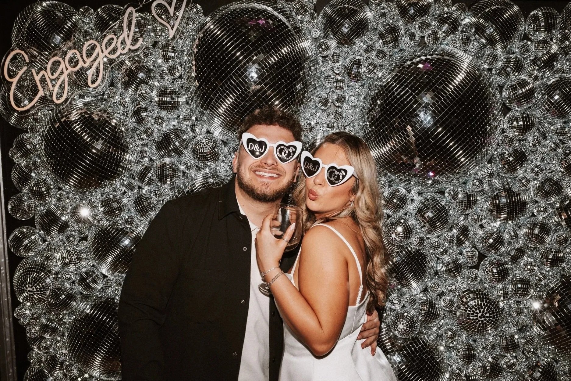 A smiling man and woman wearing D&J branded sunglasses with heart-shaped lenses, posing together at a party in front of a silver disco ball backdrop with the word "Engaged" written in cursive neon. The woman is holding a drink and making a kissing face.