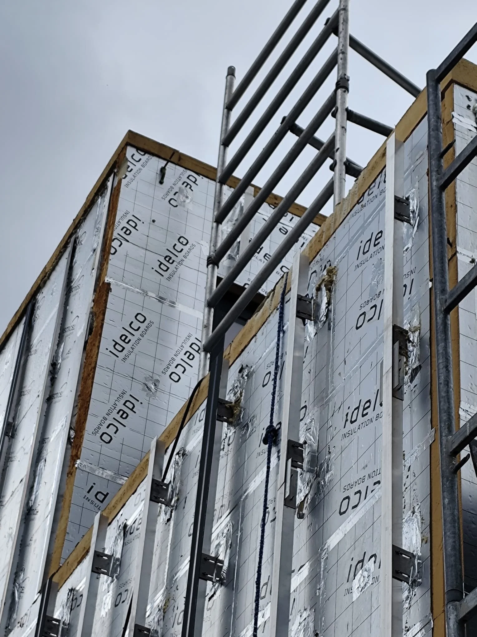 A building under construction with insulation boards labeled 'idelco' fixed on the exterior walls, and a metal scaffolding set up in front.