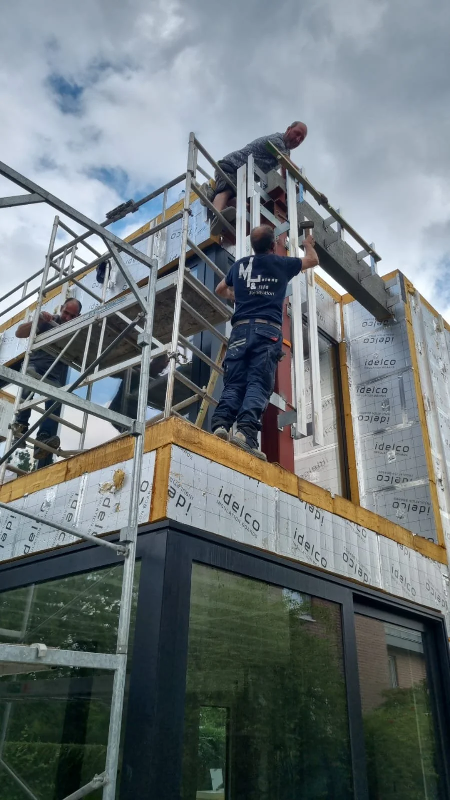 Construction workers installing exterior siding on the upper floor of a building, using scaffolding and tools, with a cloudy sky in the background.