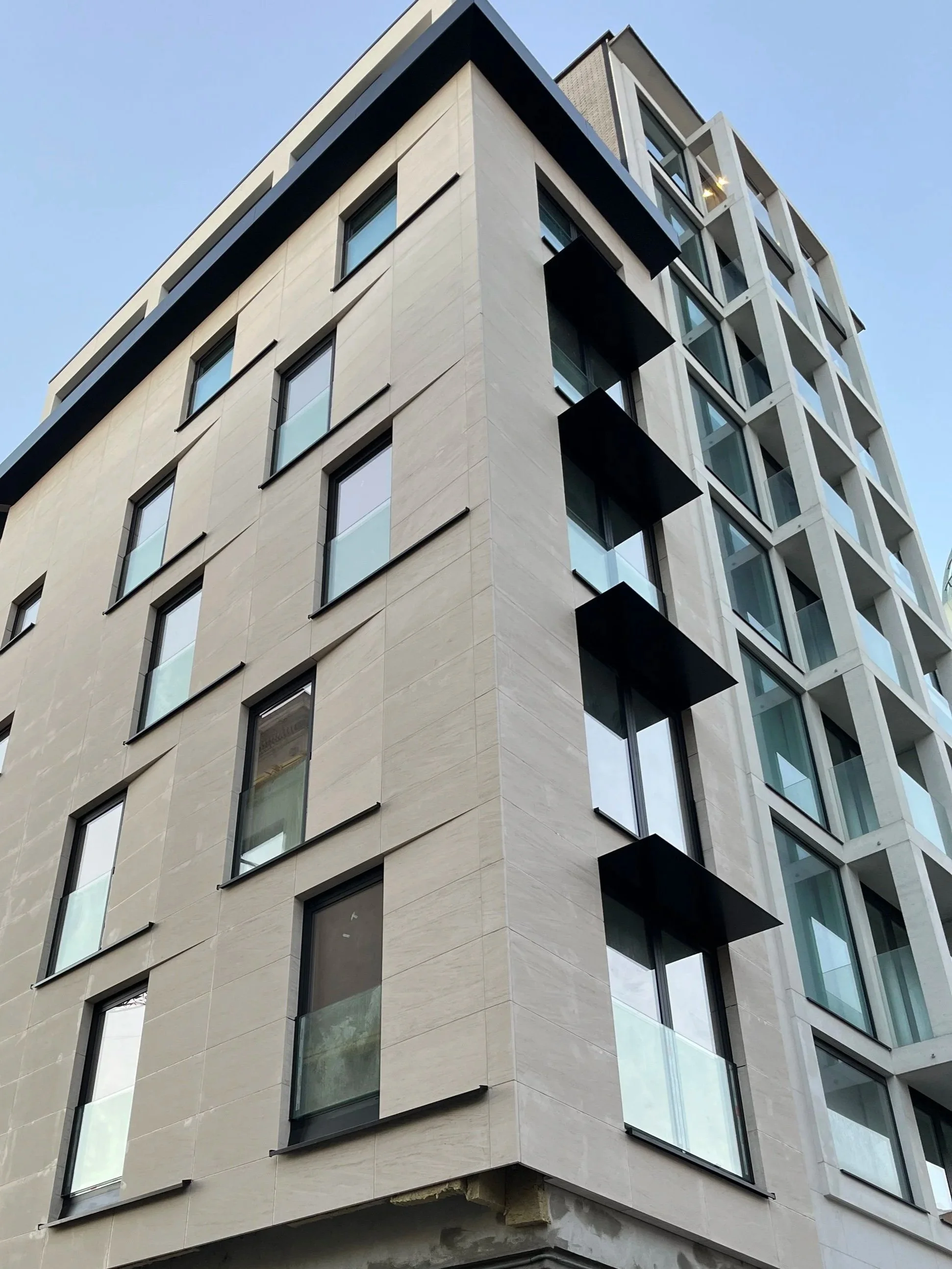 Modern multi-story building with glass windows and black balconies, viewed from below against a blue sky.