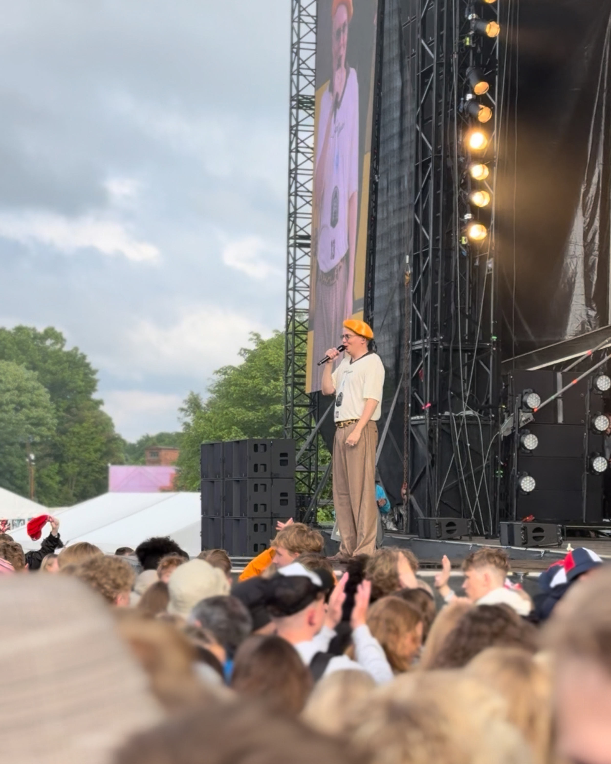 En person på scenen til en koncert, der taler eller synger i en mikrofon, med en stor skærm bagved og en stor samling mennesker foran.