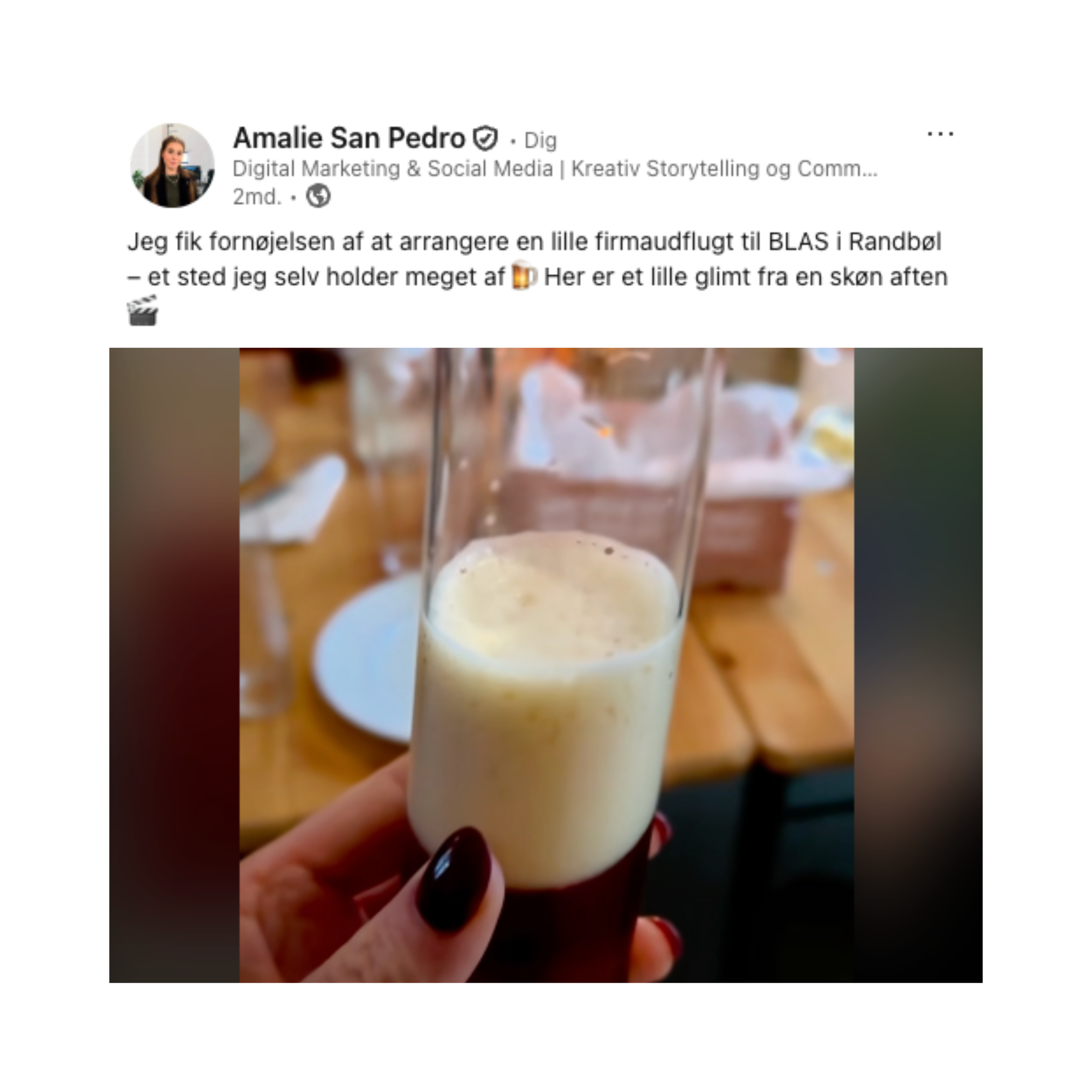 Et glas med en fyldt, lyst, skummende væske, set i en persons hånd med mørk neglelak. Baggrunden er en træbordplade med nogle fade og tallerkener, som er let slørede.