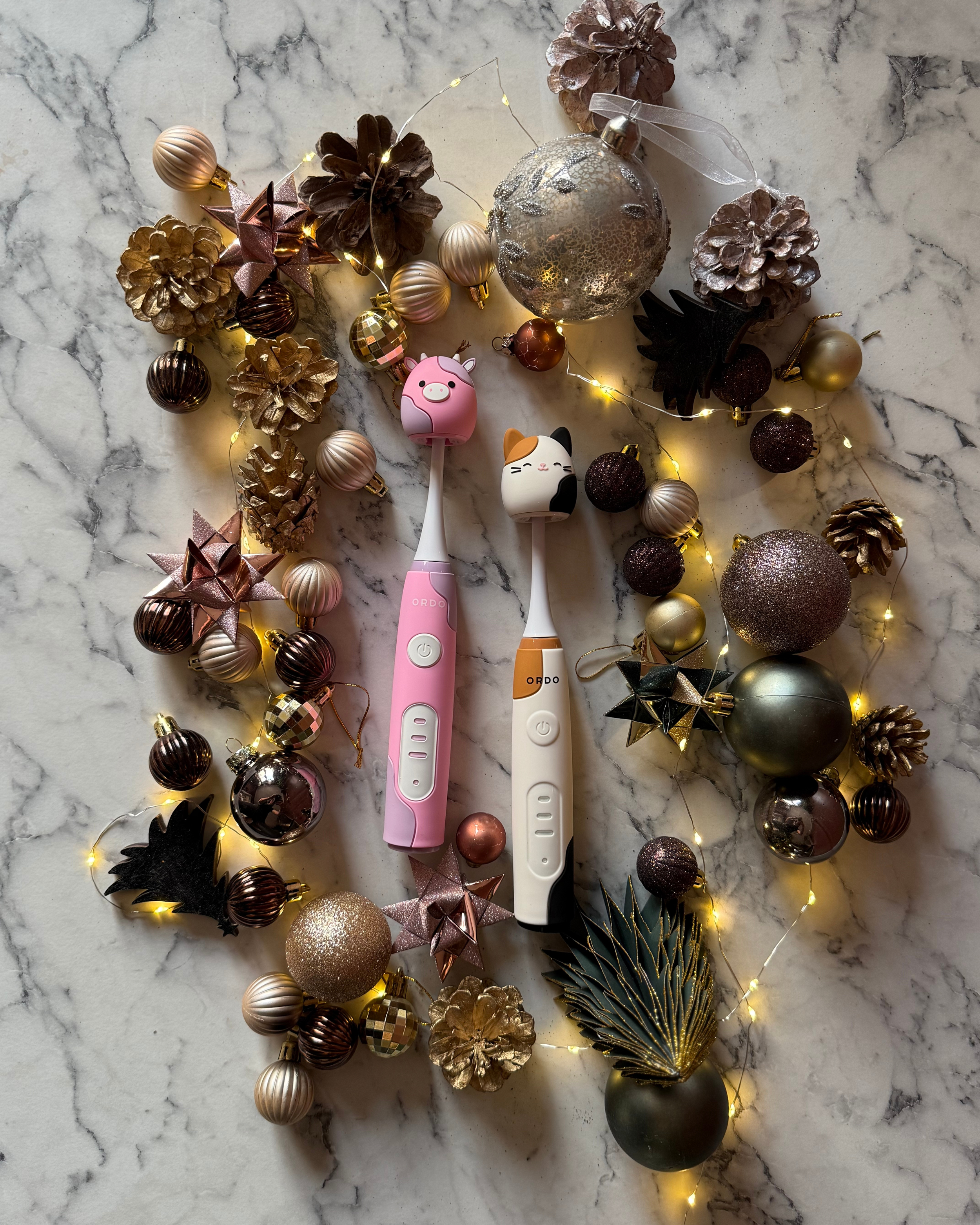 To elektriske børster med kattemotiver, omgivet af julepynt såsom gyldne, kobberfarvede og sorte julekugler, stjerner, pinecones, loofahs, og julelys på en marmormønstret baggrund.