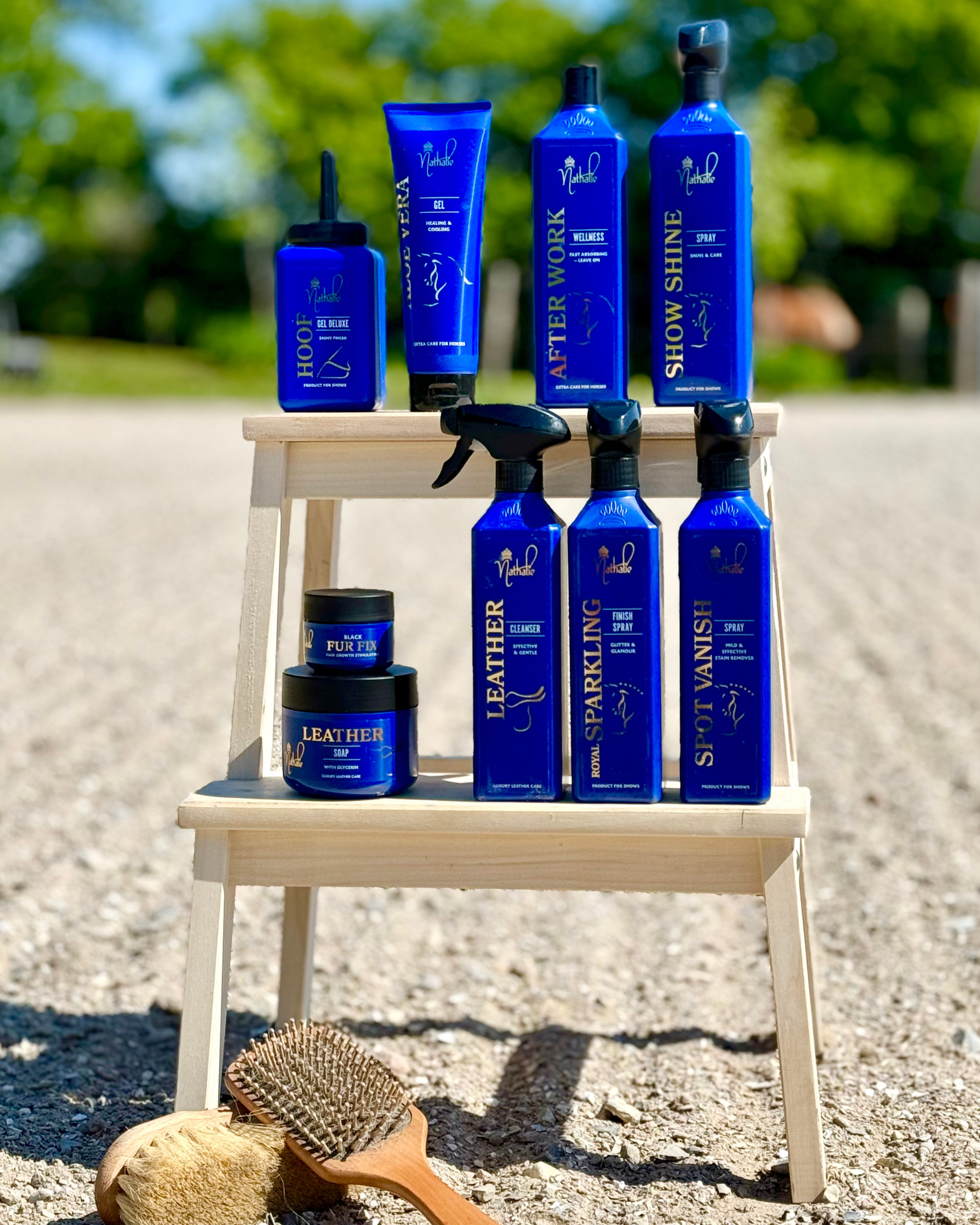 Hårplejeprodukter til heste, herunder gel, after work, shine spray, læderpleje, og plejeprodukter til læder, på en træhylde udendørs, med en børste og svamp foran.