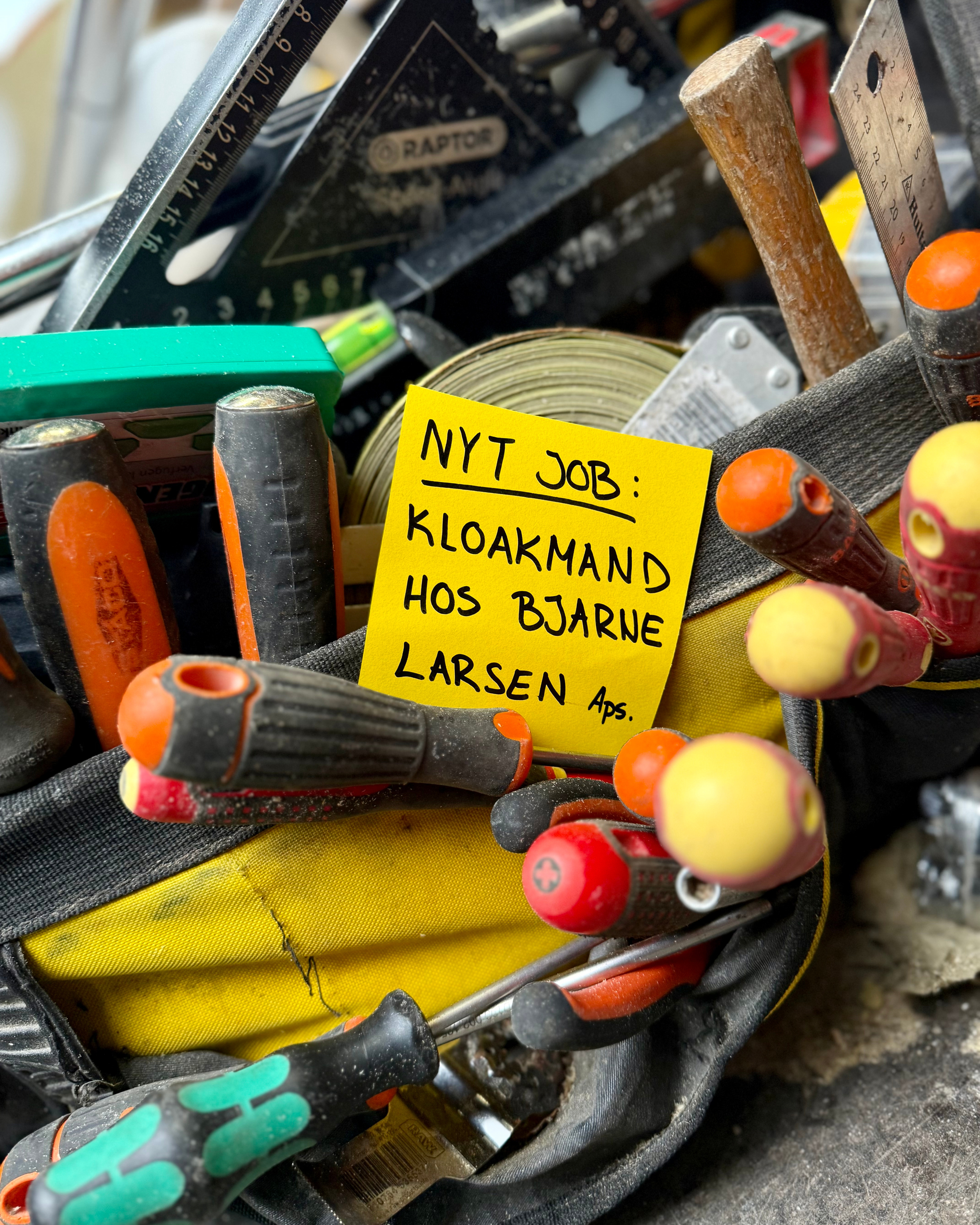 Kasse med forskellige håndværktøjer og en gul post-it note med tekst på dansk.