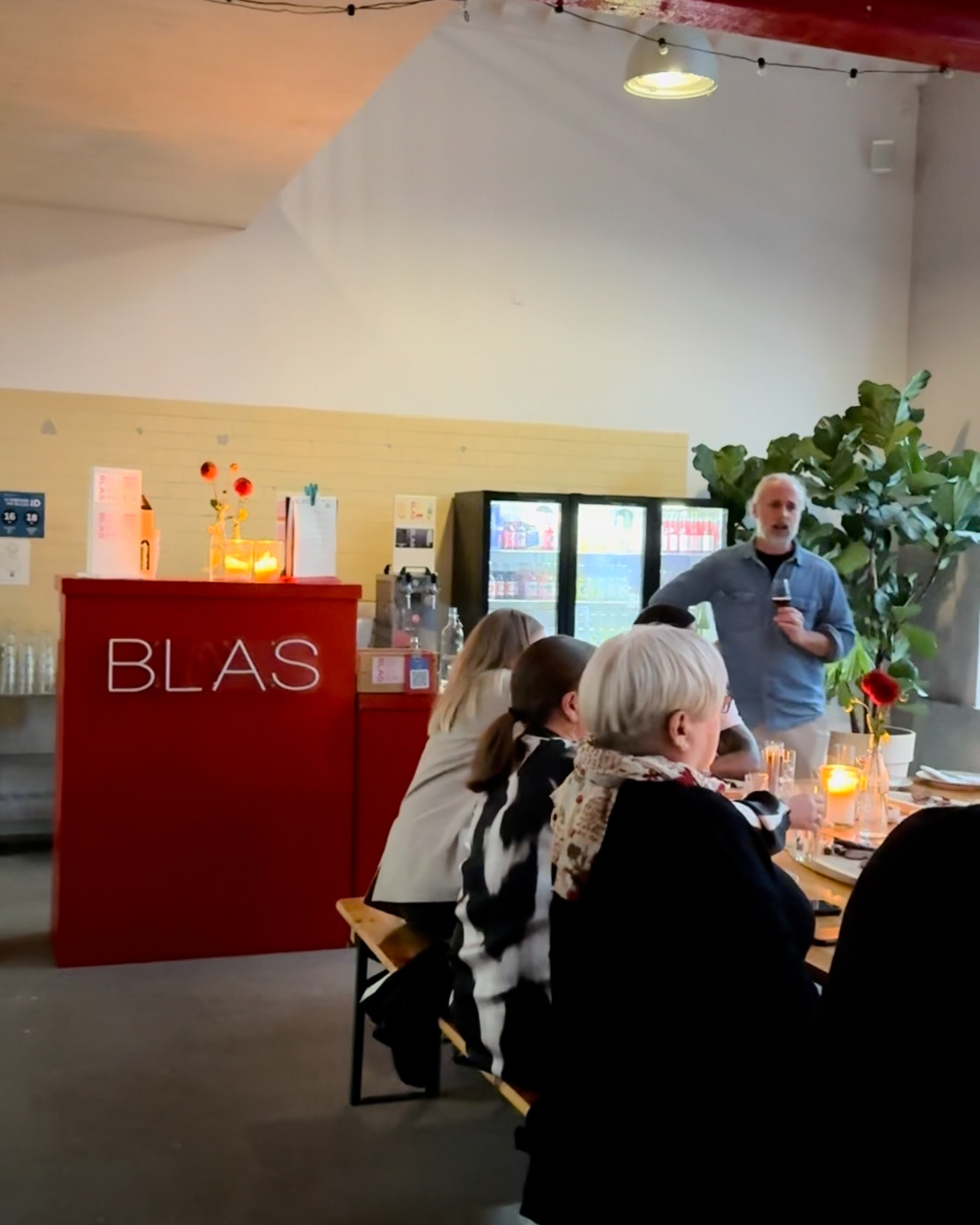 Indendørs restaurantscene med en gruppe mennesker sidder ved et langbord og lytter til en mand, der taler med en mikrofon. Bagved er et rødt barområde med 'BLÅS' skrevet på, kilde til lys og nogle planter.