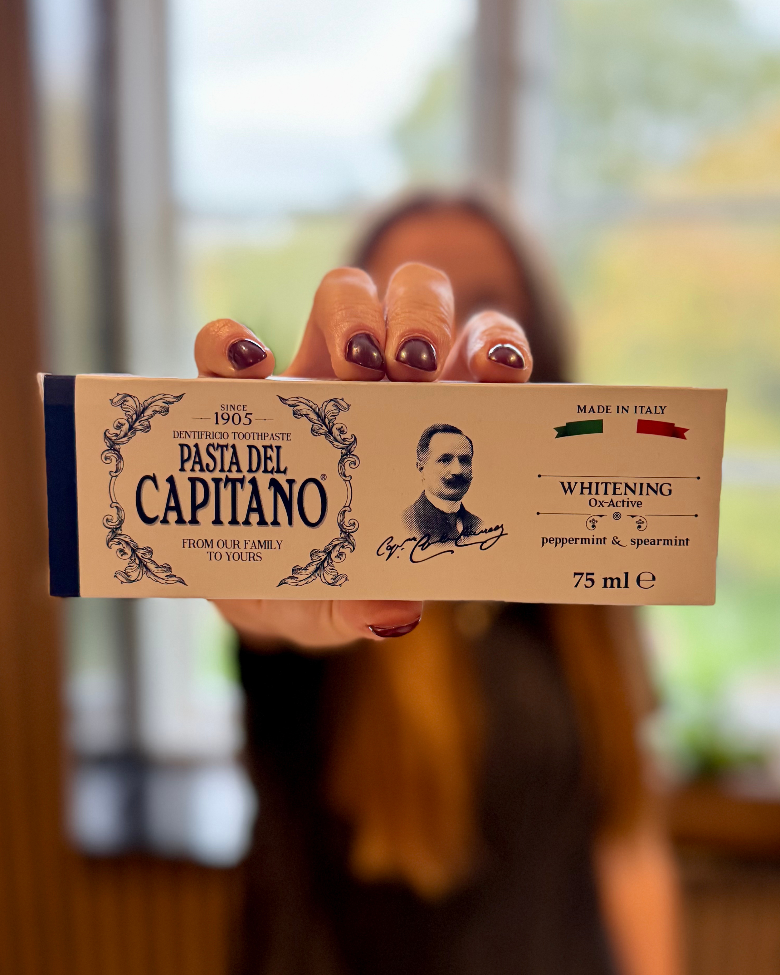 En person holder en æske tandpasta mod kameraet, ansigtet er ukendt. Æsken er fra mærket Pasta del Capitano og er i størrelsen 75 ml. Baggrunden er uklar og viser et vindue med udendørs udsigt.