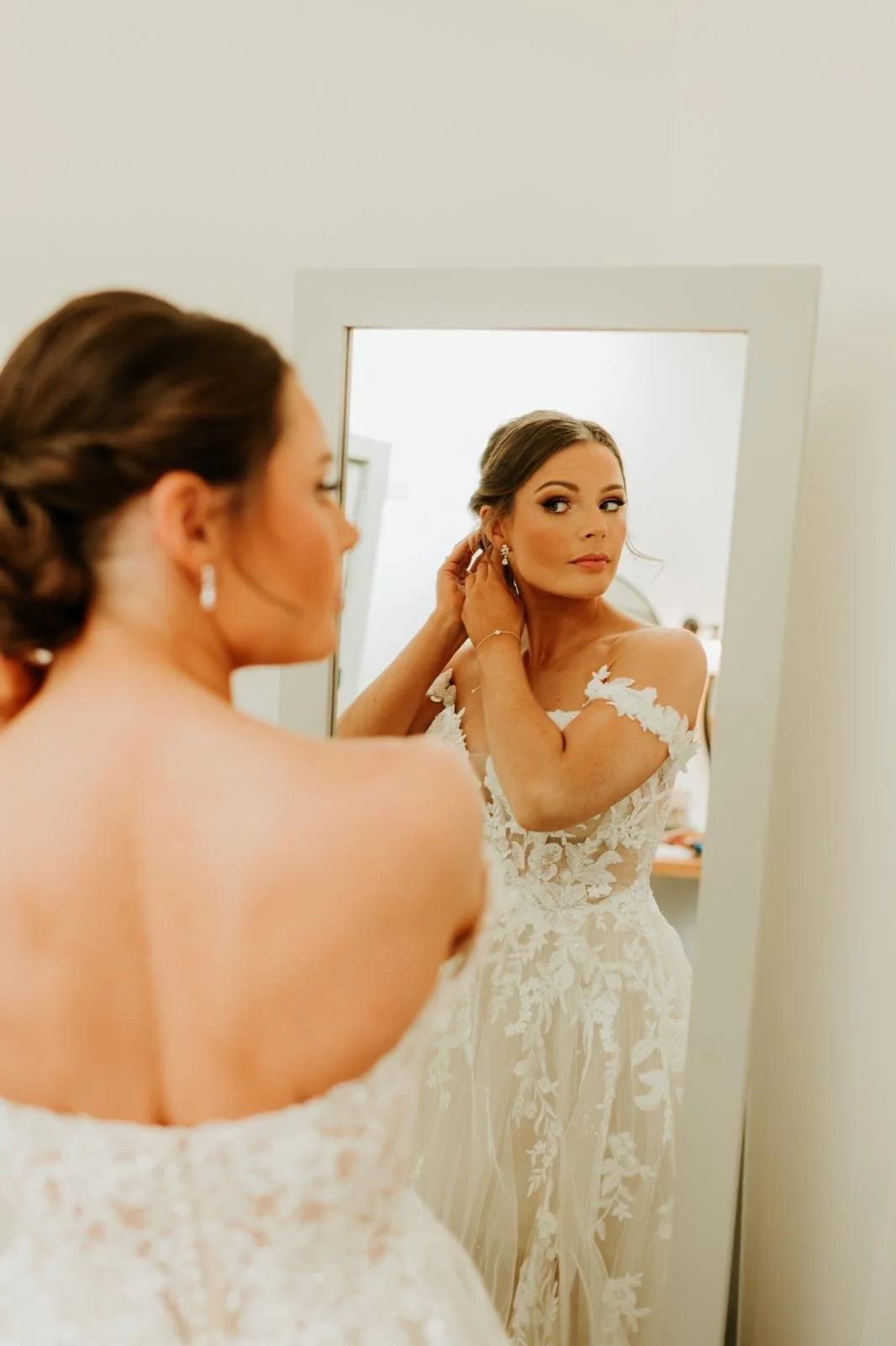 Bride in a lace wedding dress adjusting her earring while looking in a mirror.