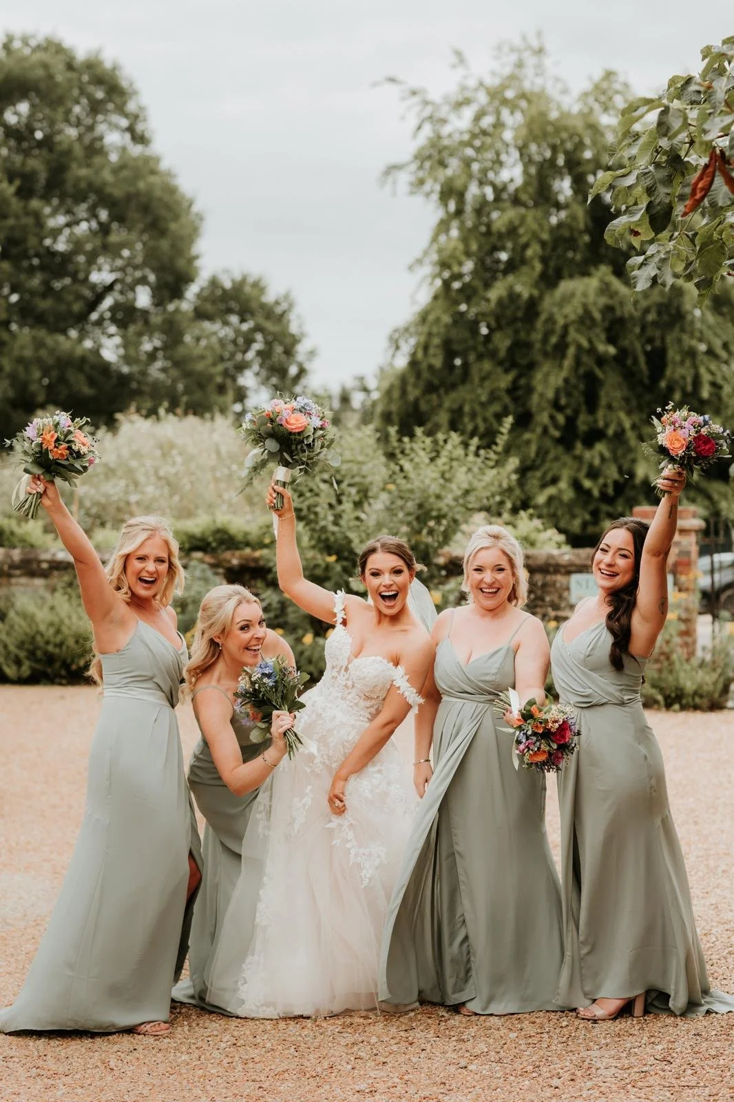 A bride and four bridesmaids celebrating outdoors, holding bouquets, with trees and a cloudy sky in the background.