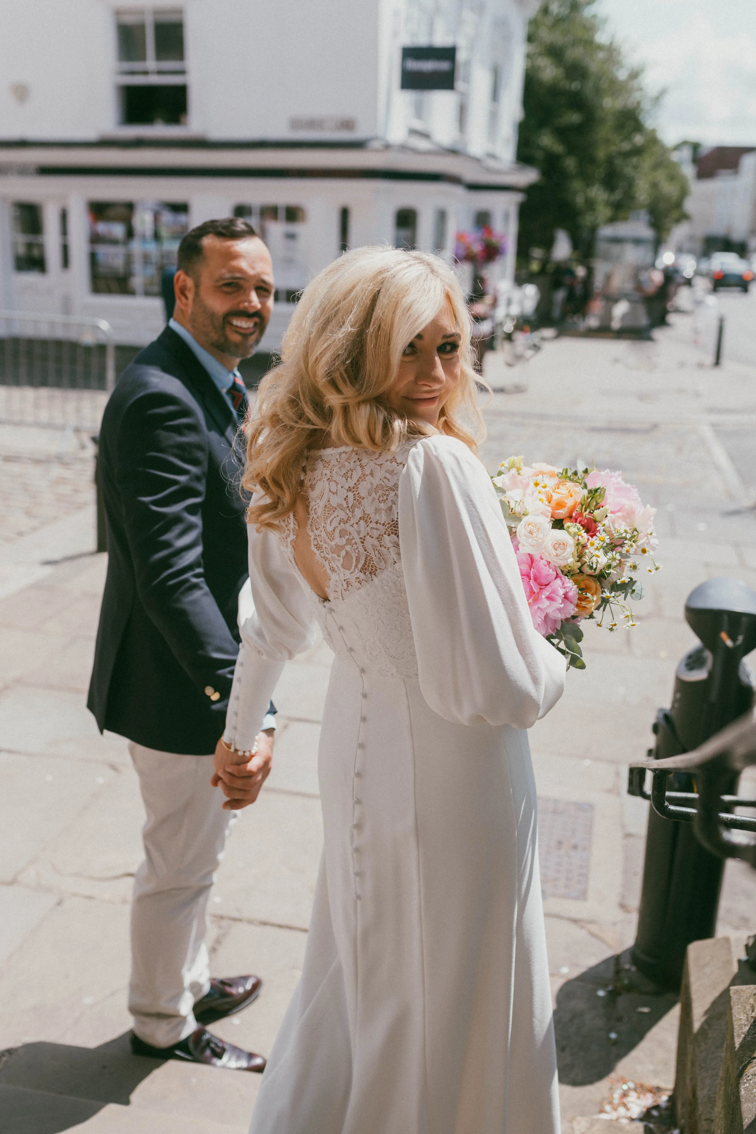 A newlywed couple holding hands outdoors, the woman in a white wedding dress holding a bouquet, the man in a dark blazer and light pants, standing on a city sidewalk with buildings and trees in the background.