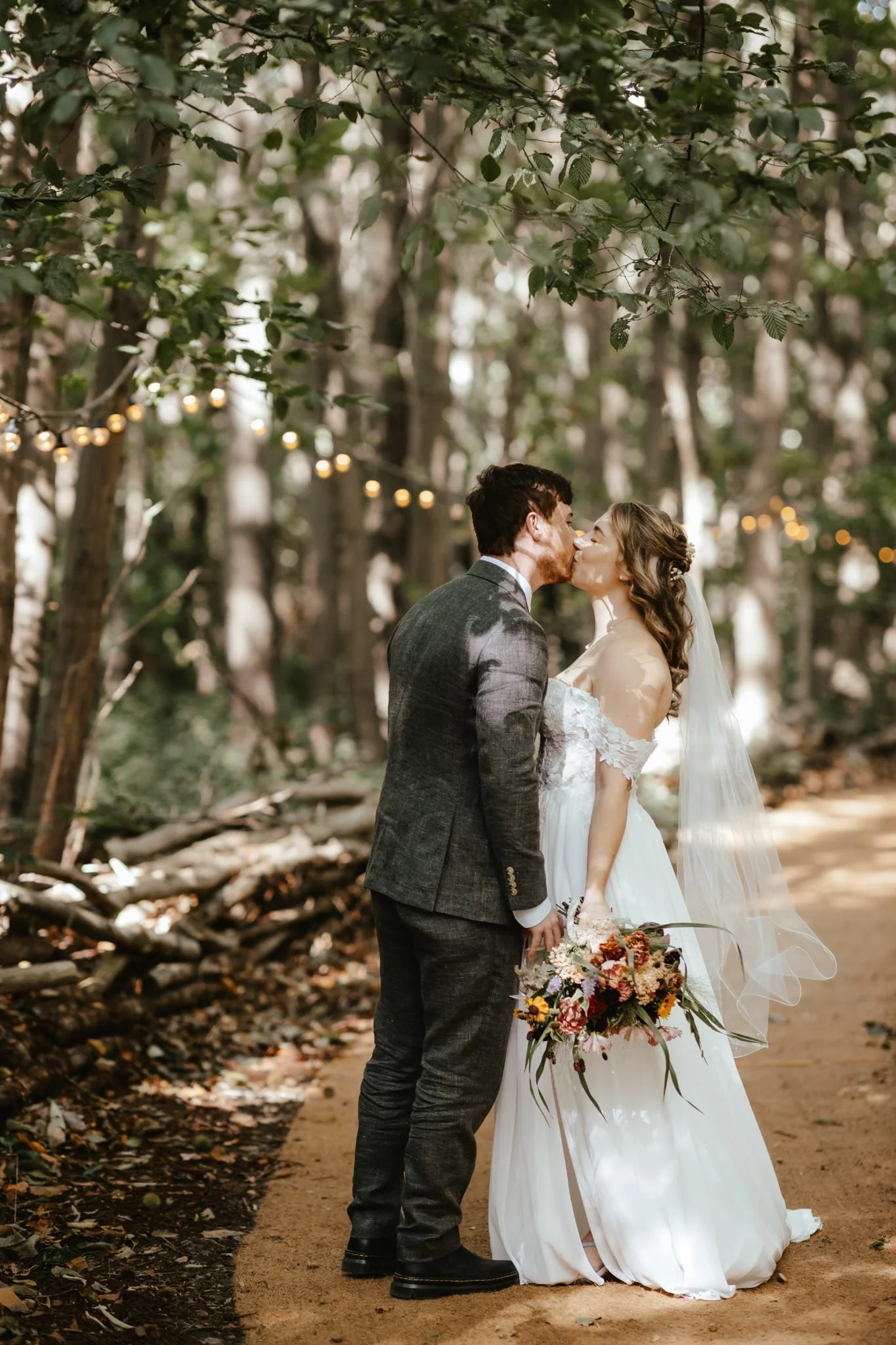 A bride and groom sharing a kiss in a wooded outdoor setting decorated with string lights, with the bride holding a bouquet of flowers.