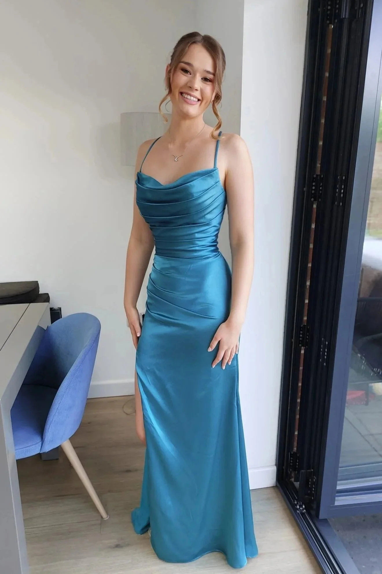 Young woman in a blue satin evening gown standing indoors next to a window.