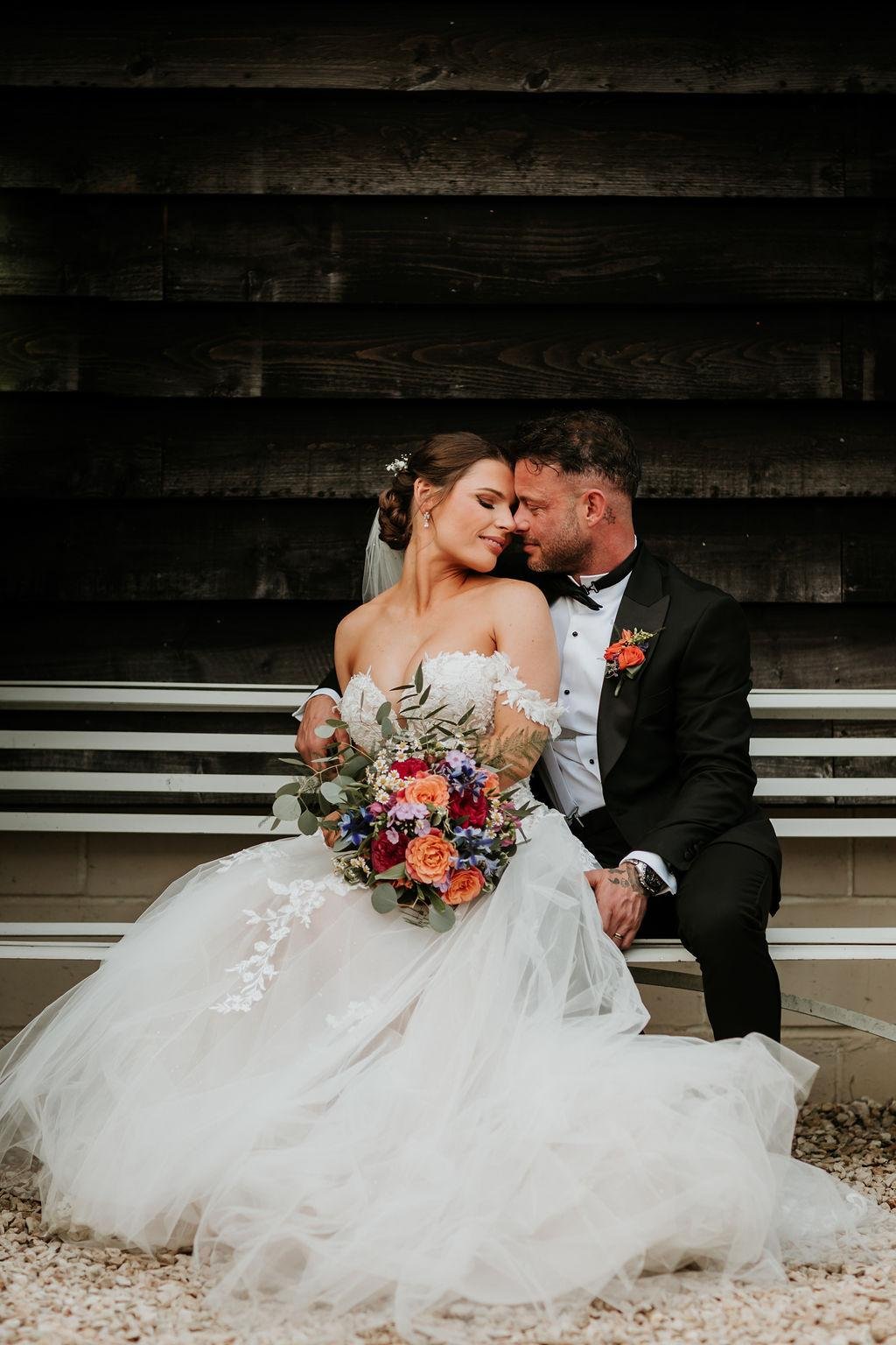 A bride and groom sitting close together on a bench, touching foreheads and smiling with their eyes closed. The bride is wearing a white wedding gown and holding a colorful bouquet, and the groom is in a black tuxedo with a boutonniere.