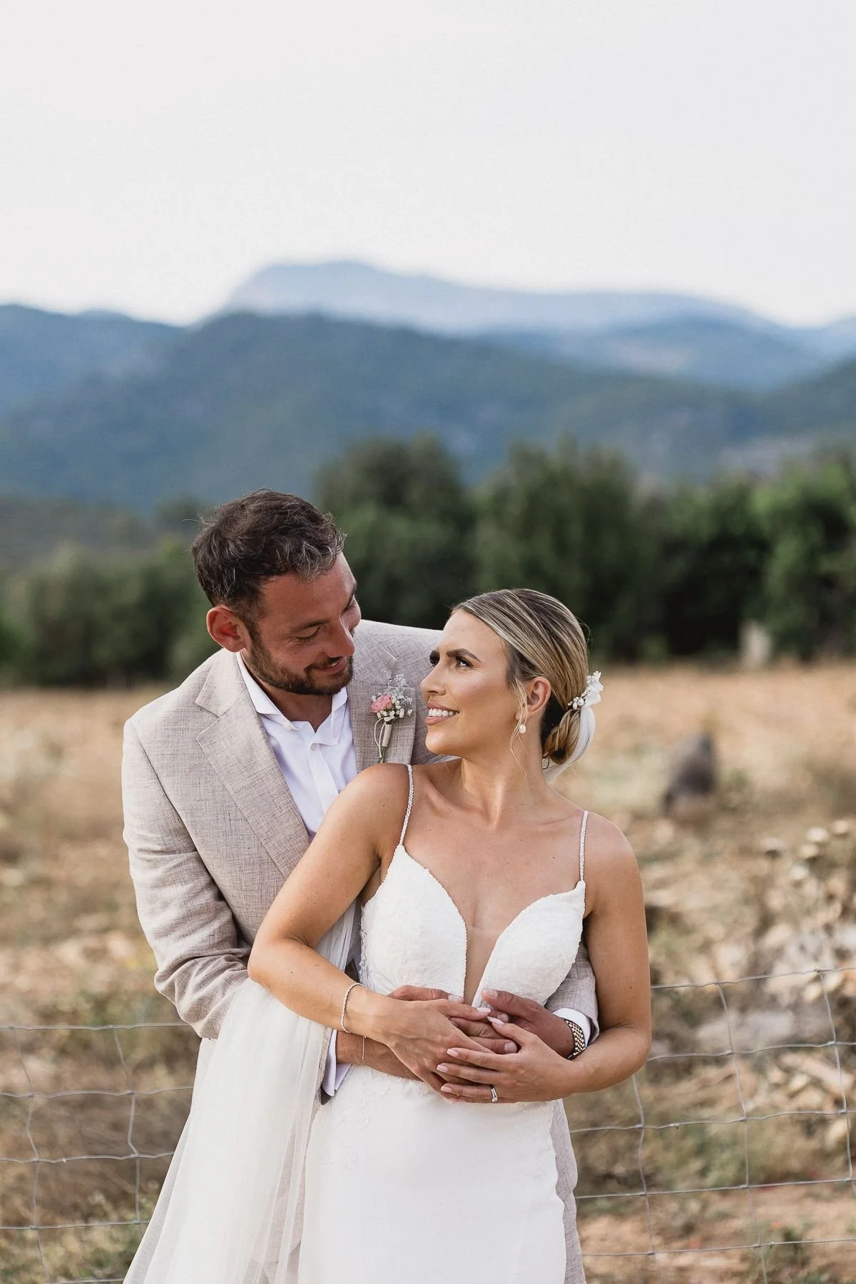 A newlywed couple stands outdoors by a wire fence, the groom in a beige suit and the bride in a white wedding dress, smiling and looking at each other with mountains and trees in the background.