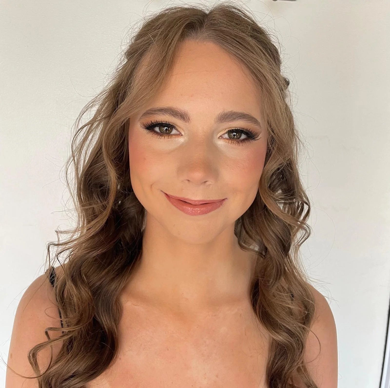 Close-up of a young woman with long, wavy brown hair and makeup, smiling and looking at the camera against a plain white background.