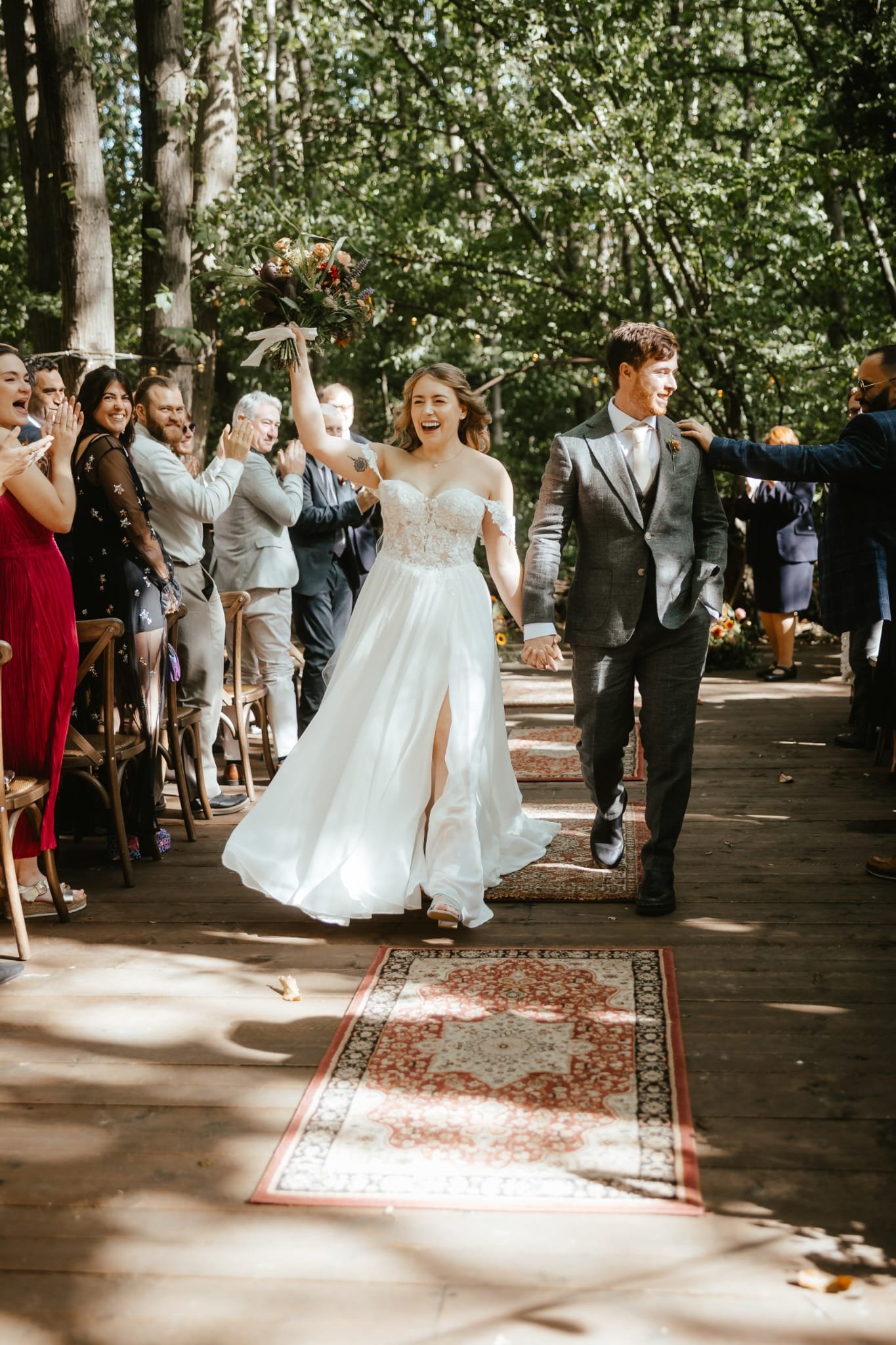 A newlywed couple walks down the aisle holding hands during an outdoor wedding ceremony, surrounded by cheerful guests in a wooded setting.