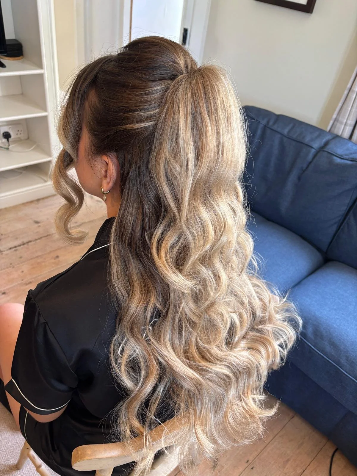 Woman with long, curly, blonde hair sitting on a chair in a room with wooden floors and a blue sofa.