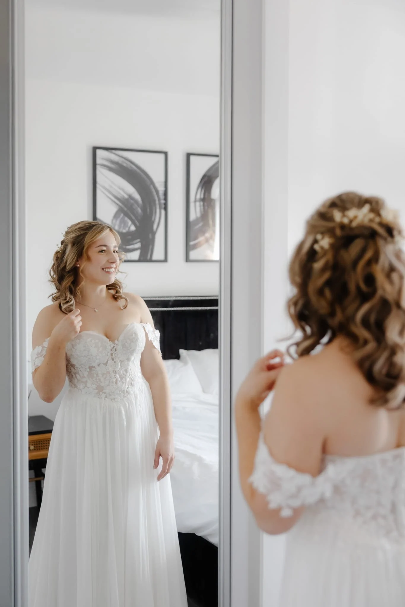 A woman in a white wedding dress looking at herself in a mirror and smiling.