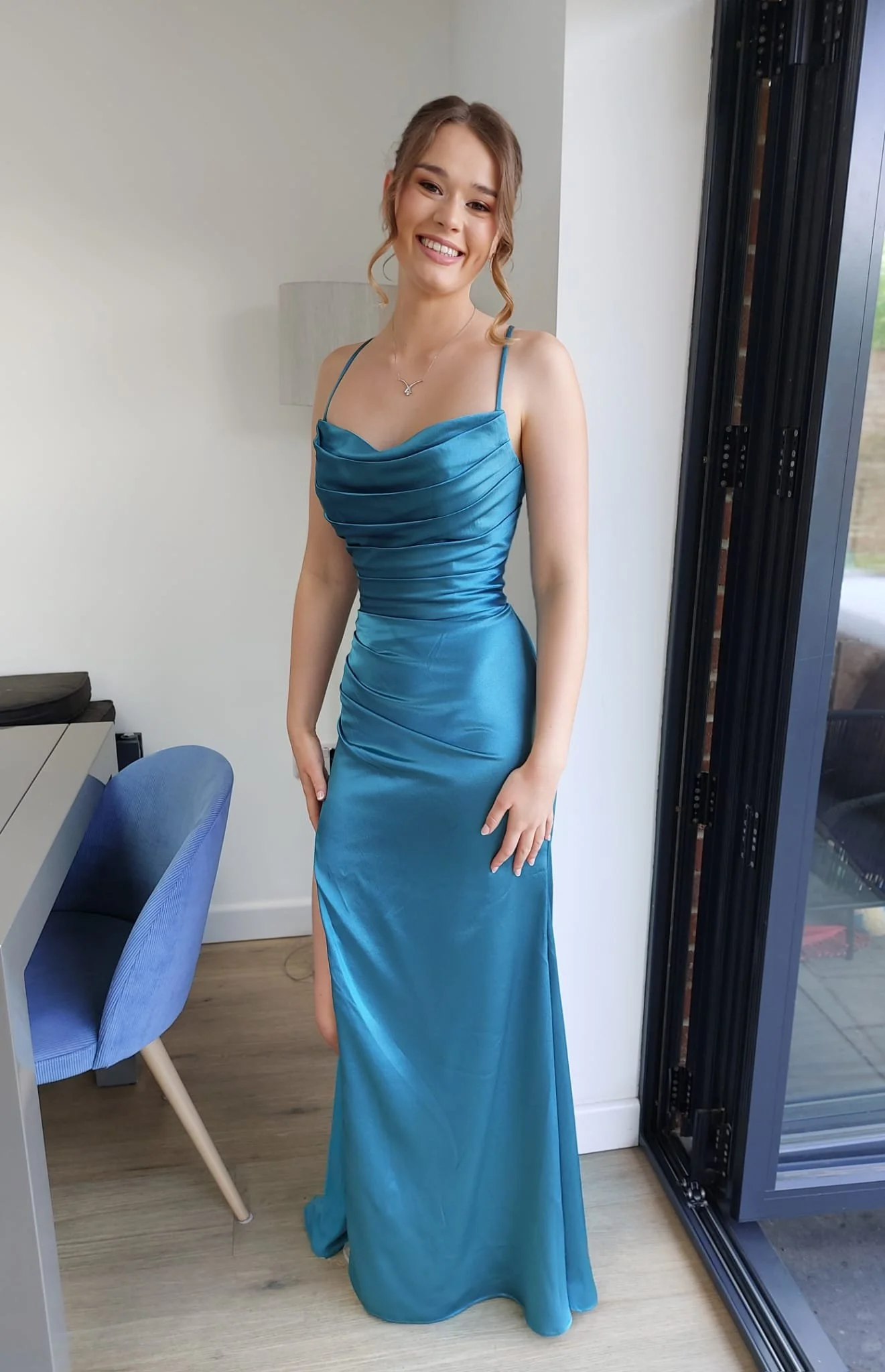 A young woman in a long, teal satin dress with spaghetti straps, standing indoors near a window, smiling at the camera.