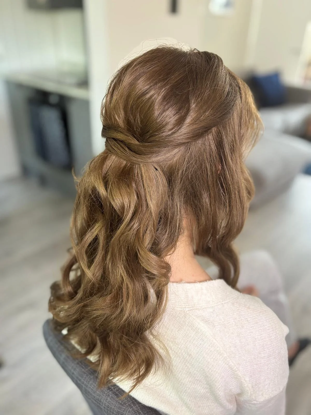 Woman with long, wavy, light brown hair styled with a twist half-updo, seated in a bright room.