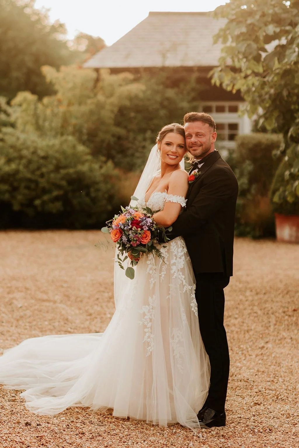 A bride and groom embrace outdoors during sunset. The bride wears a white lace wedding dress with a long train and holds a bouquet of mixed flowers. The groom is dressed in a black suit with a boutonniere, and they are smiling at the camera against a