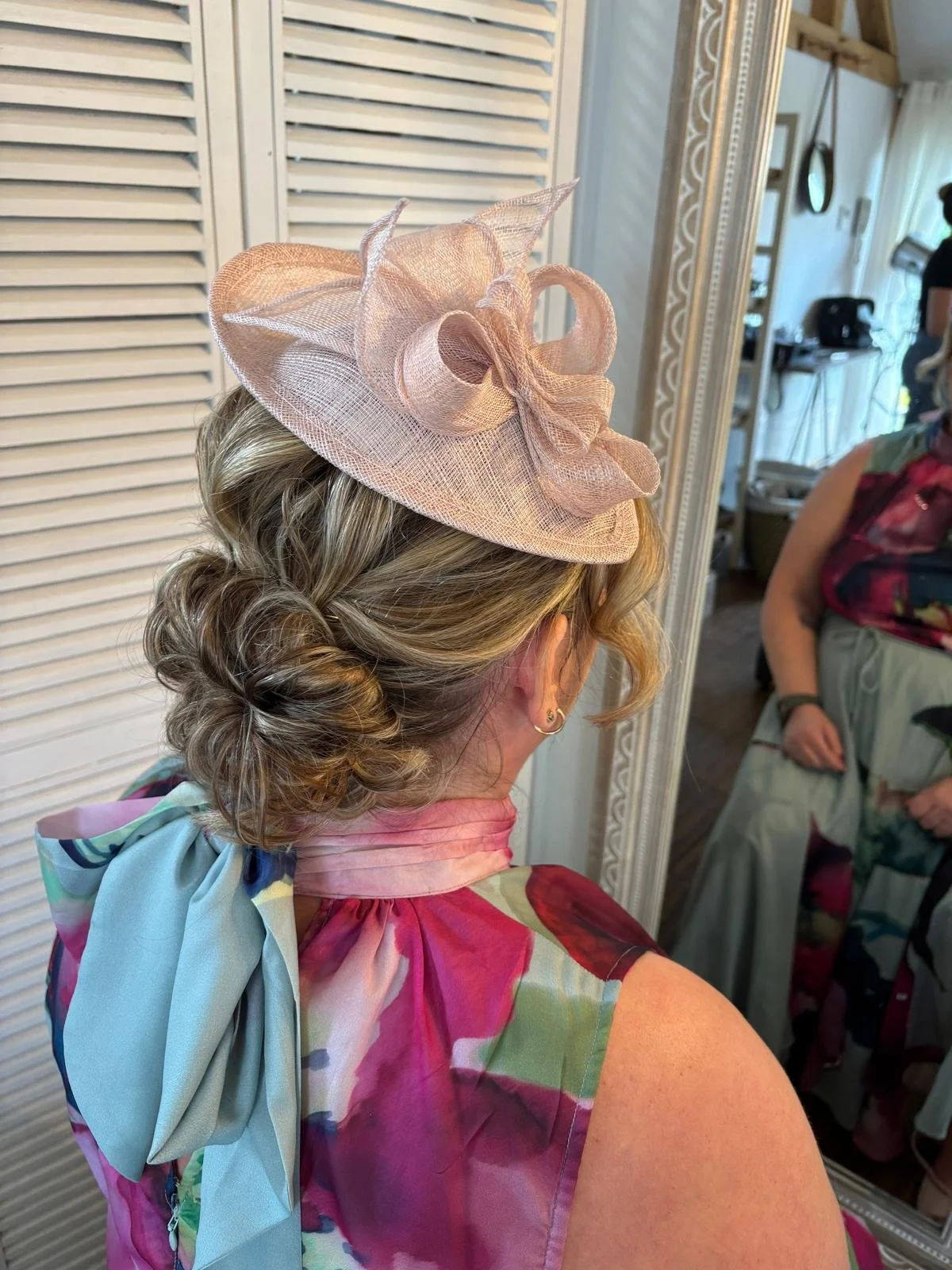 A woman with styled curly hair wearing a pink floral dress and a pink fascinator hat with decorative ribbon, standing in front of a mirror.