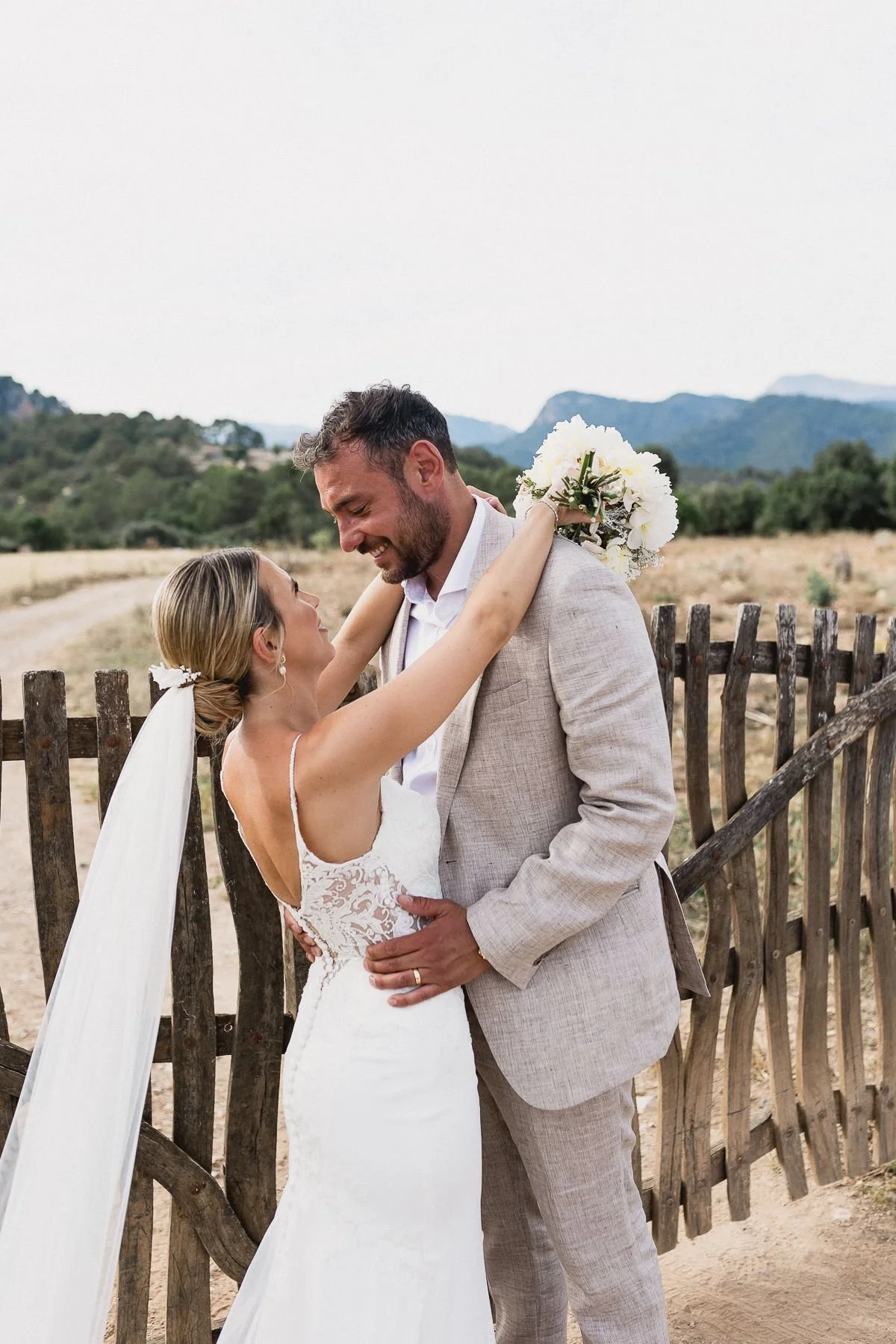 A bride and groom embrace outdoors, smiling at each other, with the bride raising a bouquet of white flowers. The background features a rustic wooden fence, open fields, trees, and distant mountains.