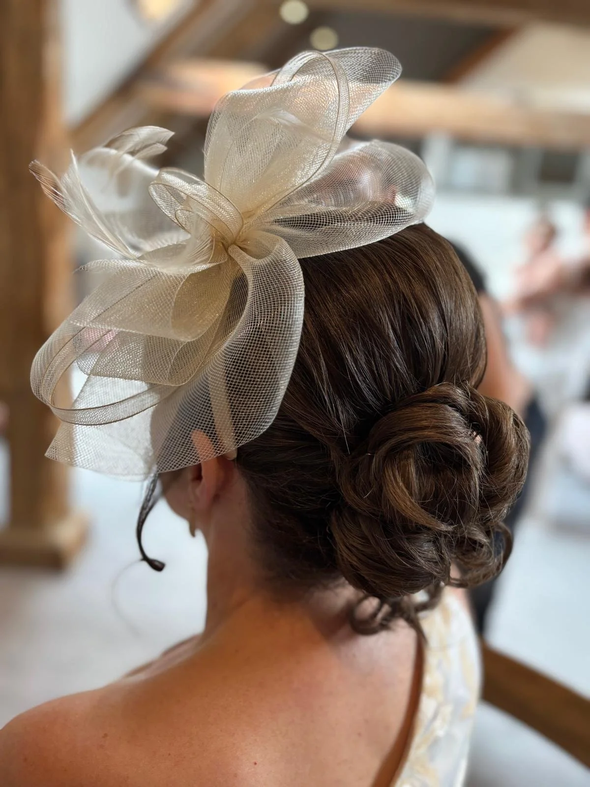 Back view of a woman with elegant brown hairstyle pinned up, wearing a beige floral dress and a large cream-colored fascinator with ribbon and mesh details.