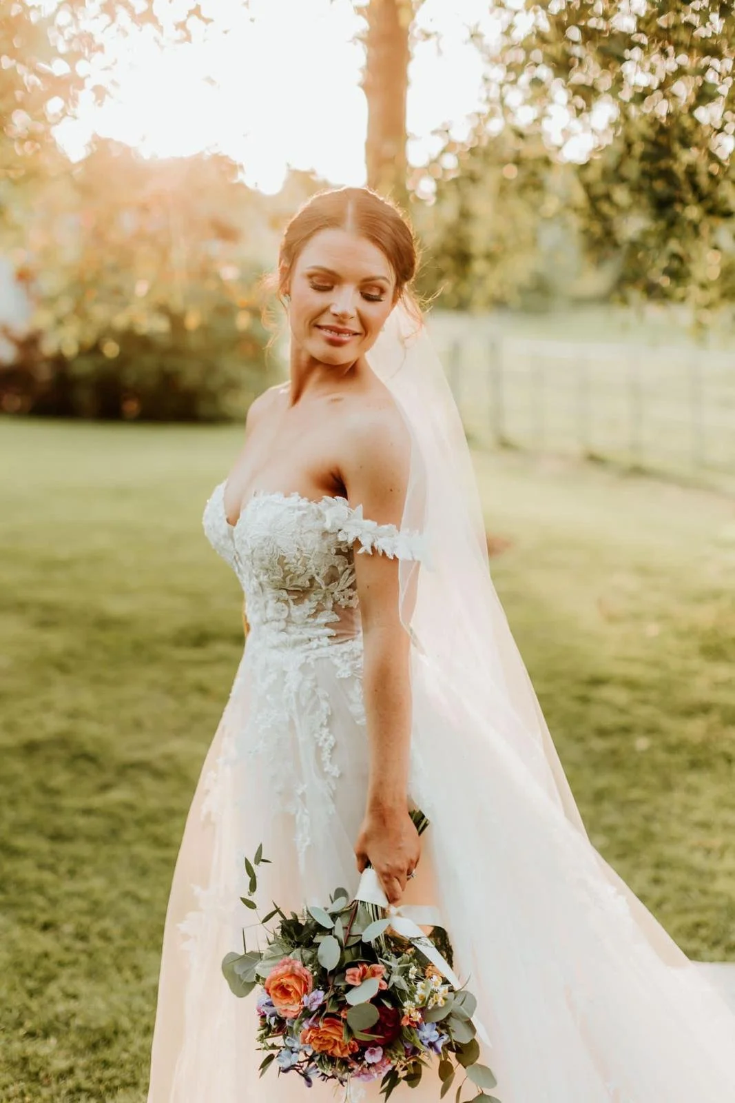 A bride in a white wedding dress with lace details, holding a colorful bouquet of roses and greenery, standing outdoors during sunset with sunlight filtering through trees.
