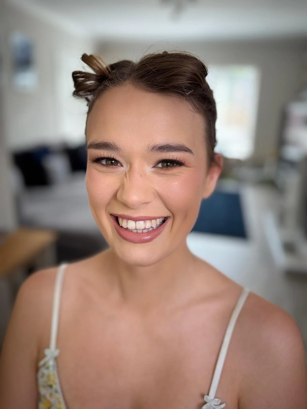 A young woman with short brown hair styled in buns, smiling broadly showing her teeth, in a bright indoor setting.
