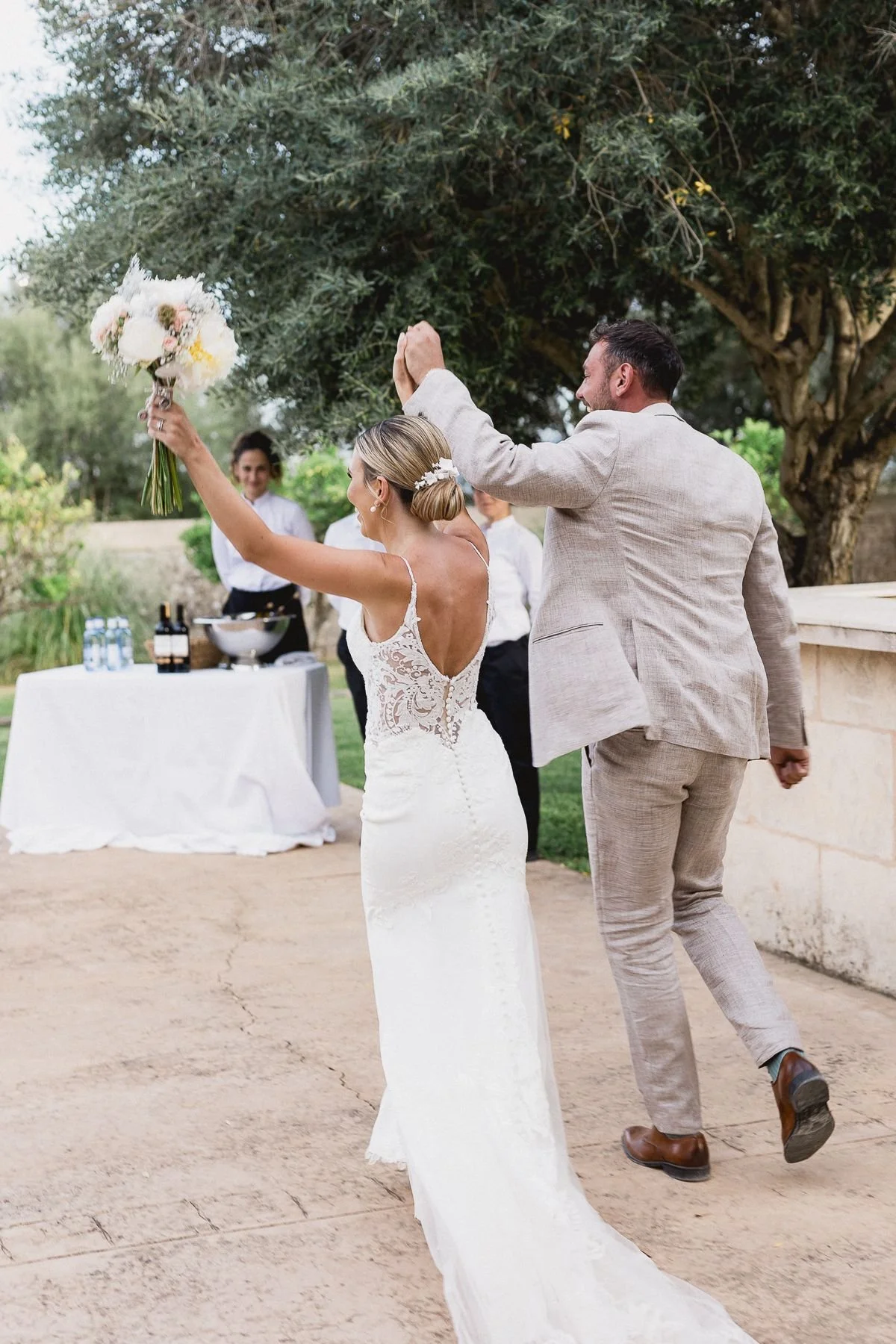 Bride in a lace wedding dress and groom in a light-colored suit celebrating outdoors with friends, holding a bouquet of flowers.