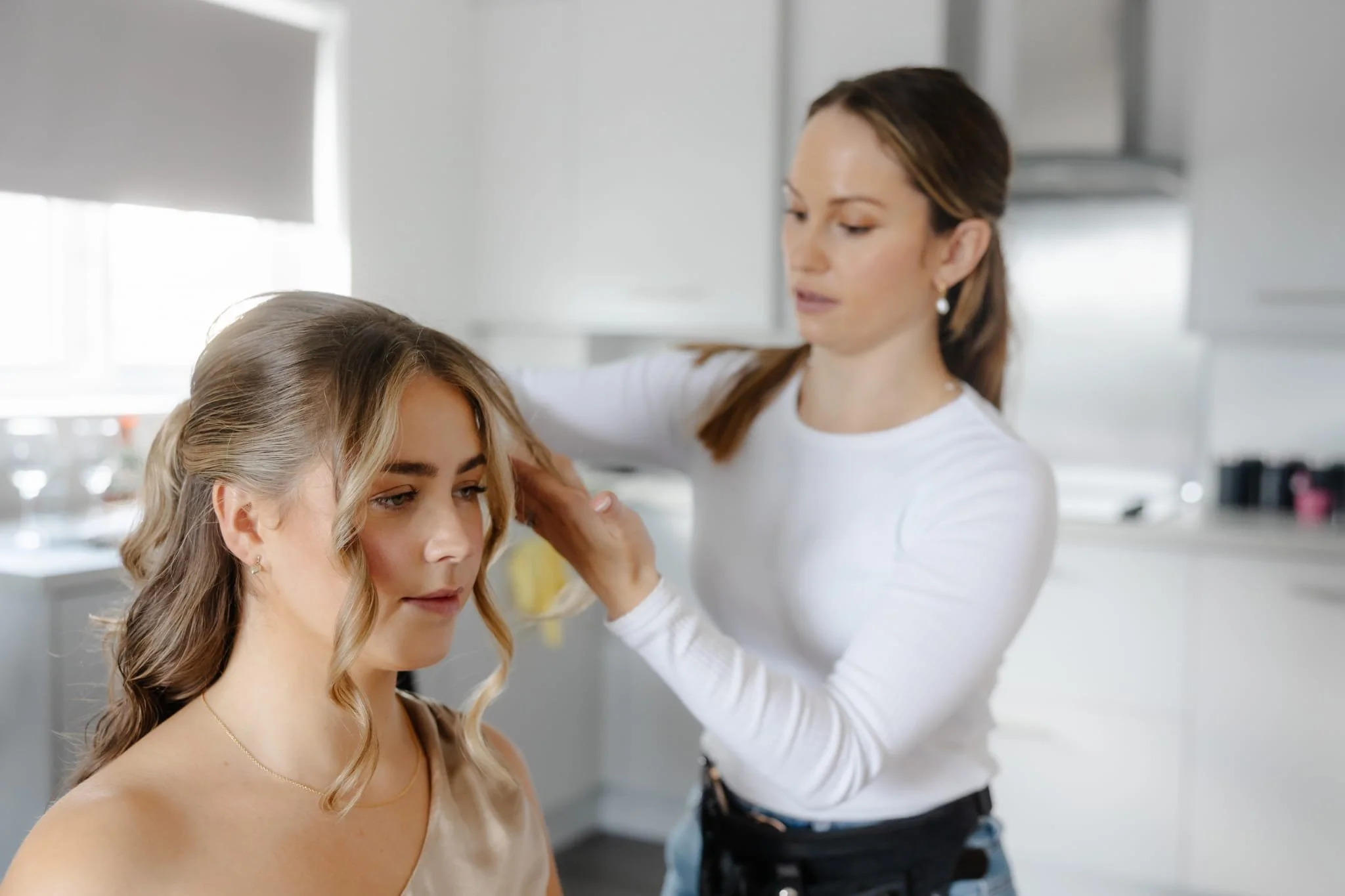 A woman with styled blonde hair sitting in a chair, getting her hair done by a stylist in a white long sleeve shirt in a bright, modern kitchen.