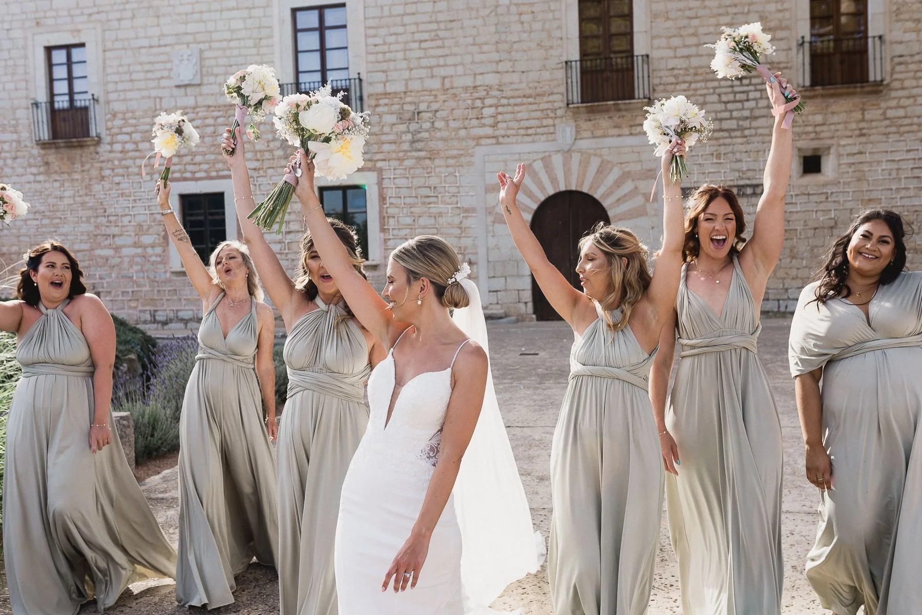 A bride and five bridesmaids outdoors celebrating, with the bridesmaids holding bouquets of flowers raised in the air, in front of a stone building with arched doorway.