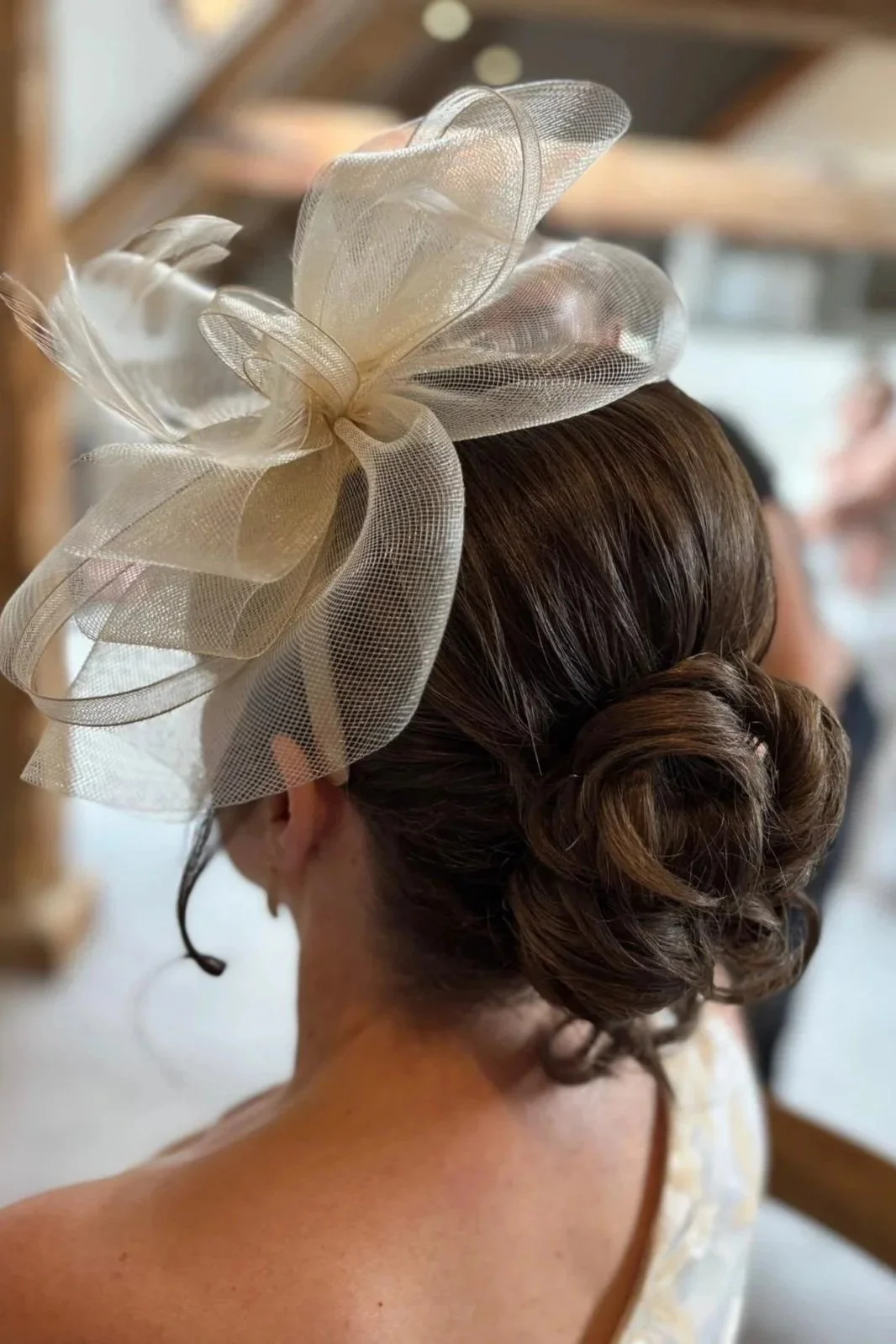 Back view of a woman with her hair styled in an elegant updo, adorned with a large, cream-colored mesh fascinator featuring layered loops and a bow.