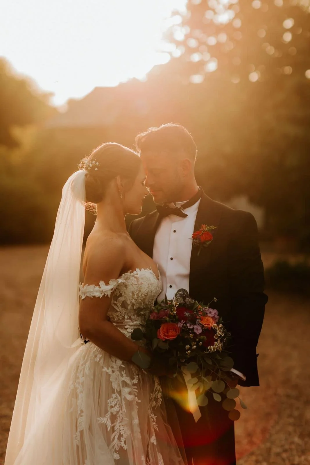 Bride and groom in wedding attire sharing an intimate moment outdoors at sunset, with the bride holding a bouquet of flowers.