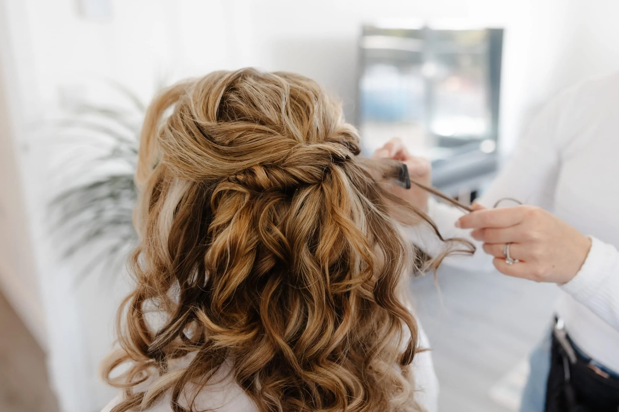 A woman with long, curly, blonde hair is getting her hair styled by a professional. The stylist is applying hair product or styling tools to her hair, which has a braid incorporated into the hairstyle. The setting appears to be a bright, modern salon