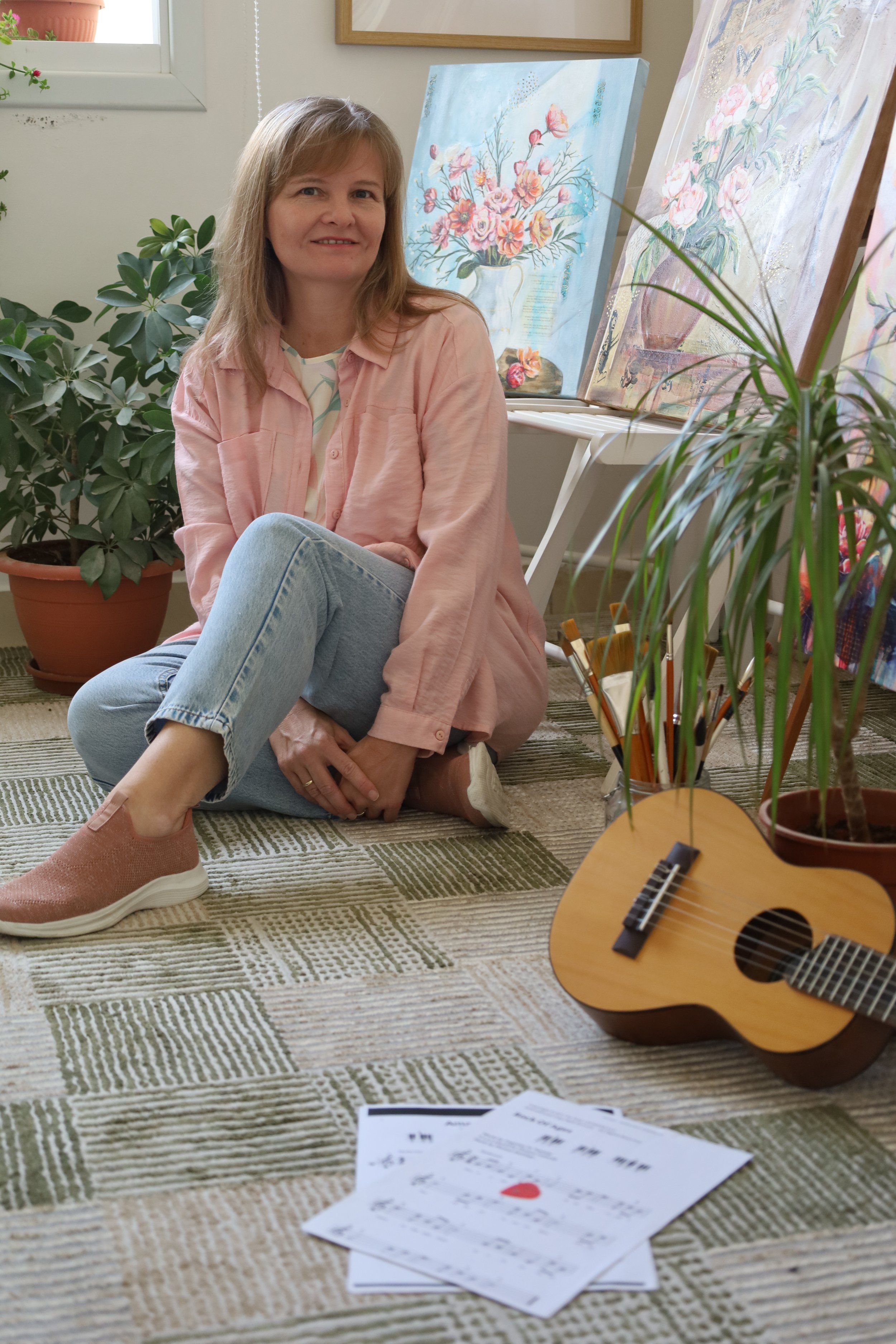 Liezle Wilken, a mixed media artist, pictured in her creative space with floral artworks. The scene includes a guitar and greenery, reflecting the ‘delicate dialogue’ and spirit of praise that inspires her work.