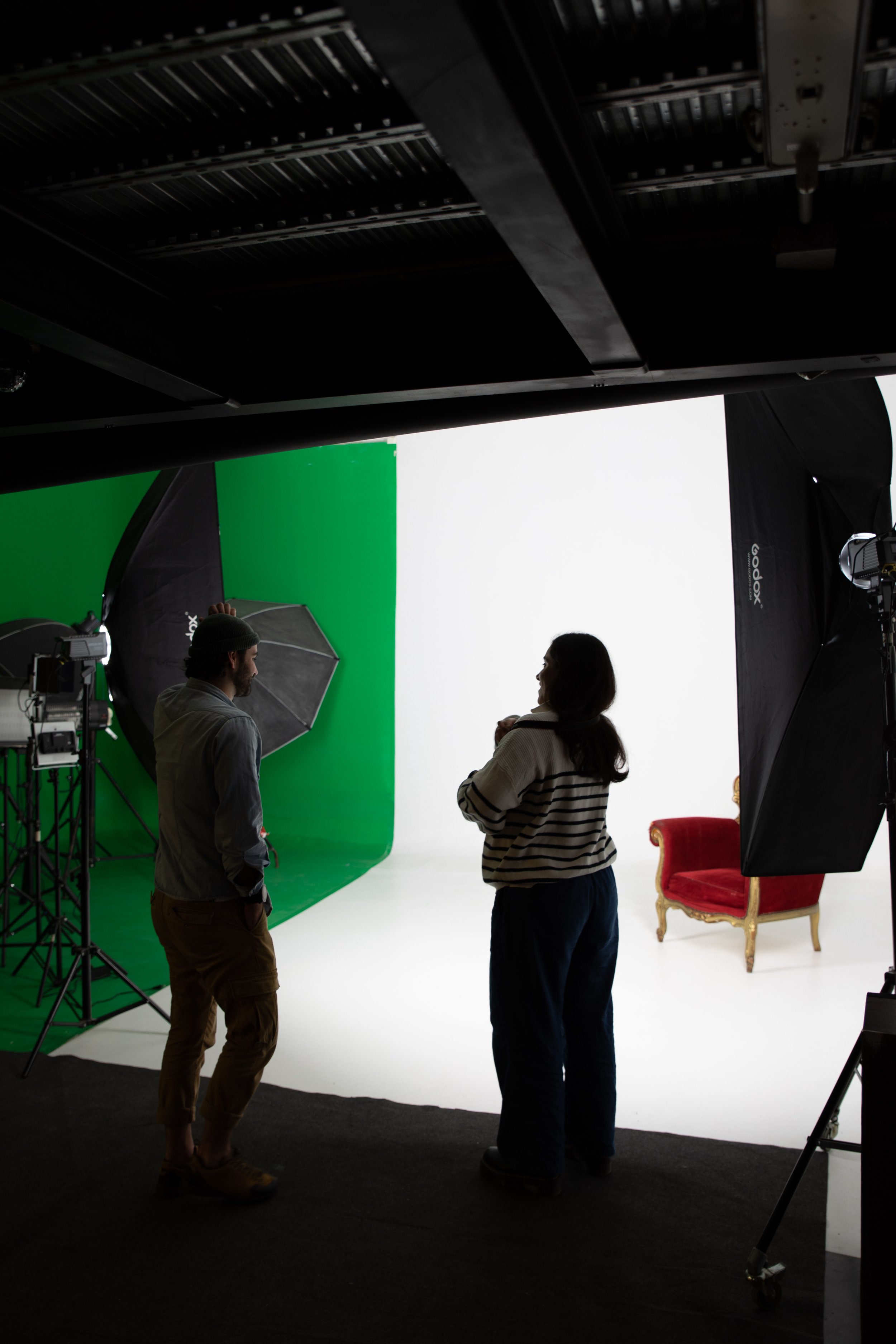 Deux personnes discutent dans un studio de tournage avec un fond vert, des équipements d'éclairage, un sofa rouge et une chaise en bois avec coussin rouge, et un ciel noir au plafond.